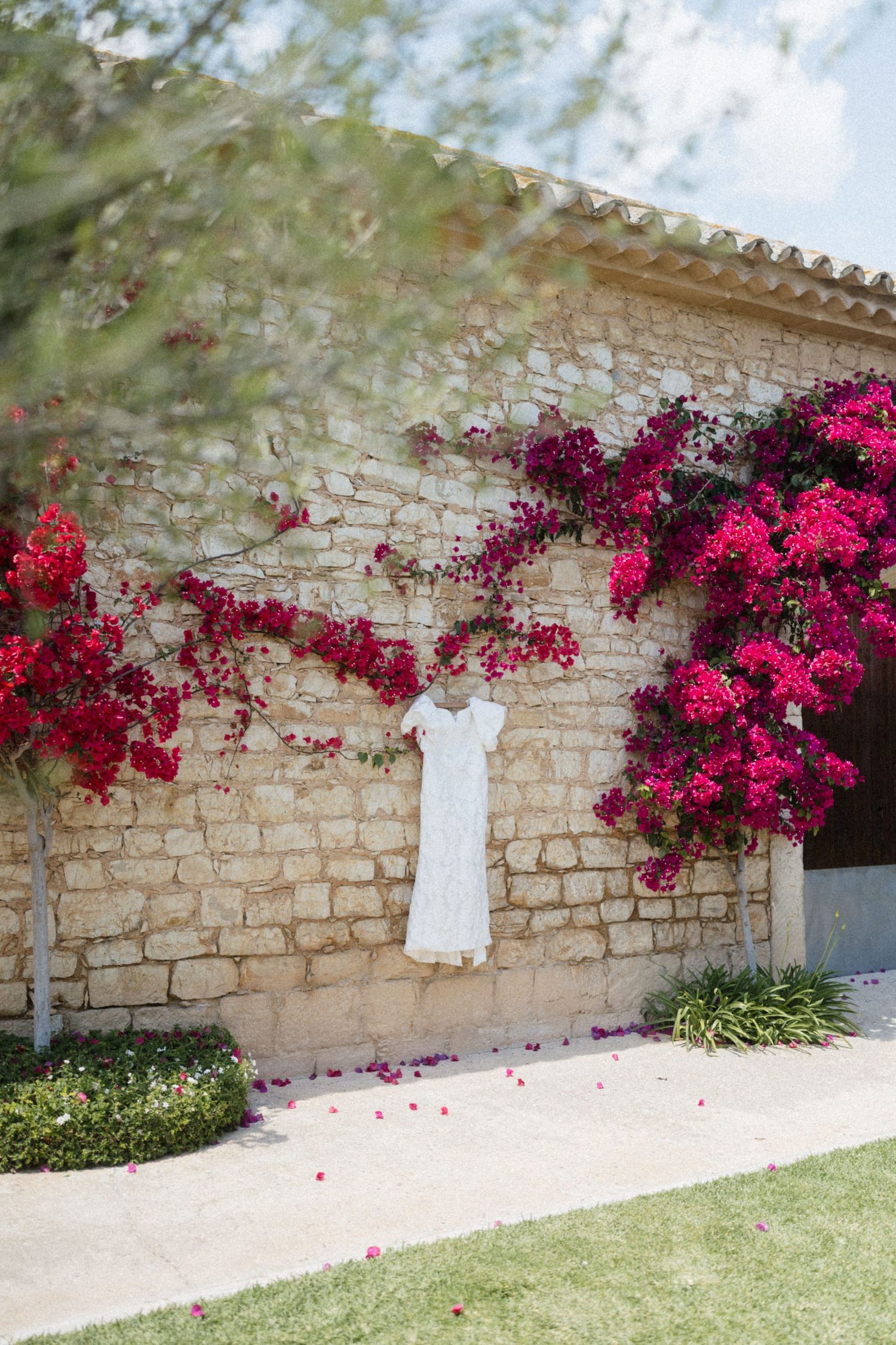 White wedding dress hanging on a stone wall adorned with vibrant pink bougainvillea flowers.