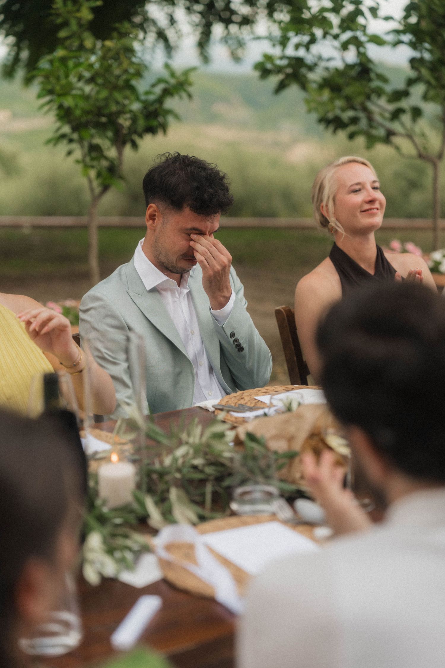 Emotional groom in sage suit wipes tears while smiling blonde guest laughs at outdoor wedding reception.