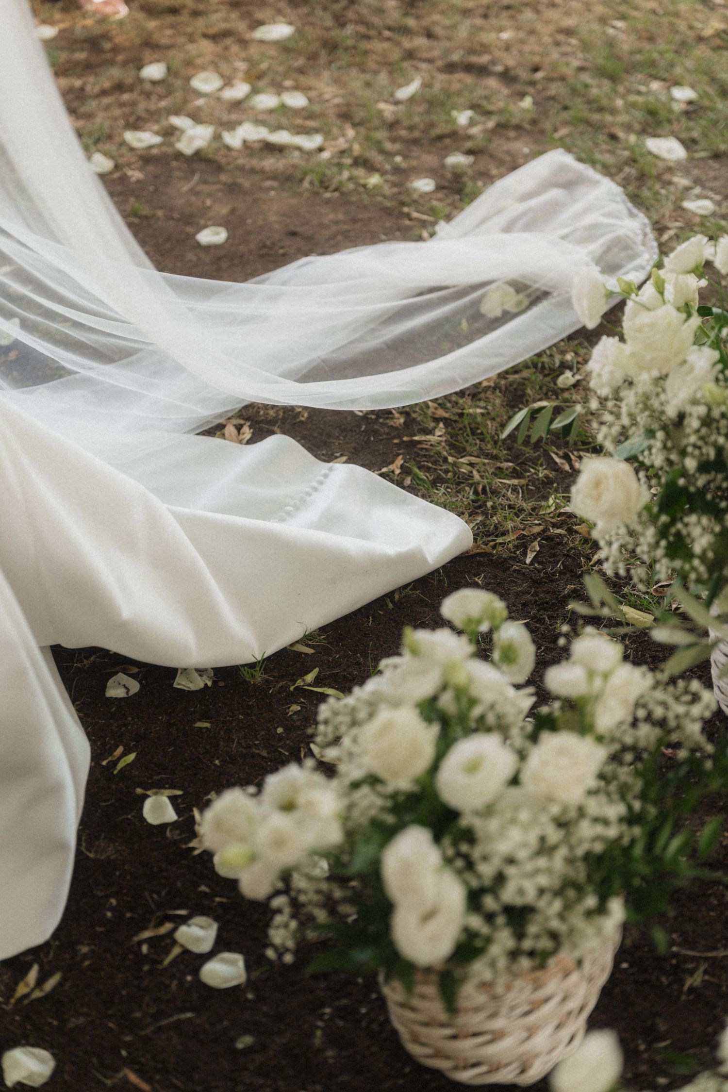 White wedding veil and dress train flowing beside basket of white roses on grass.