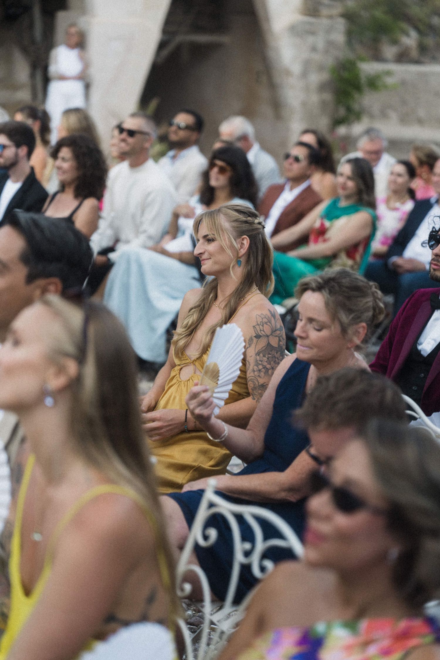 Wedding guests seated outdoors, woman in yellow dress with tattoos fans herself.