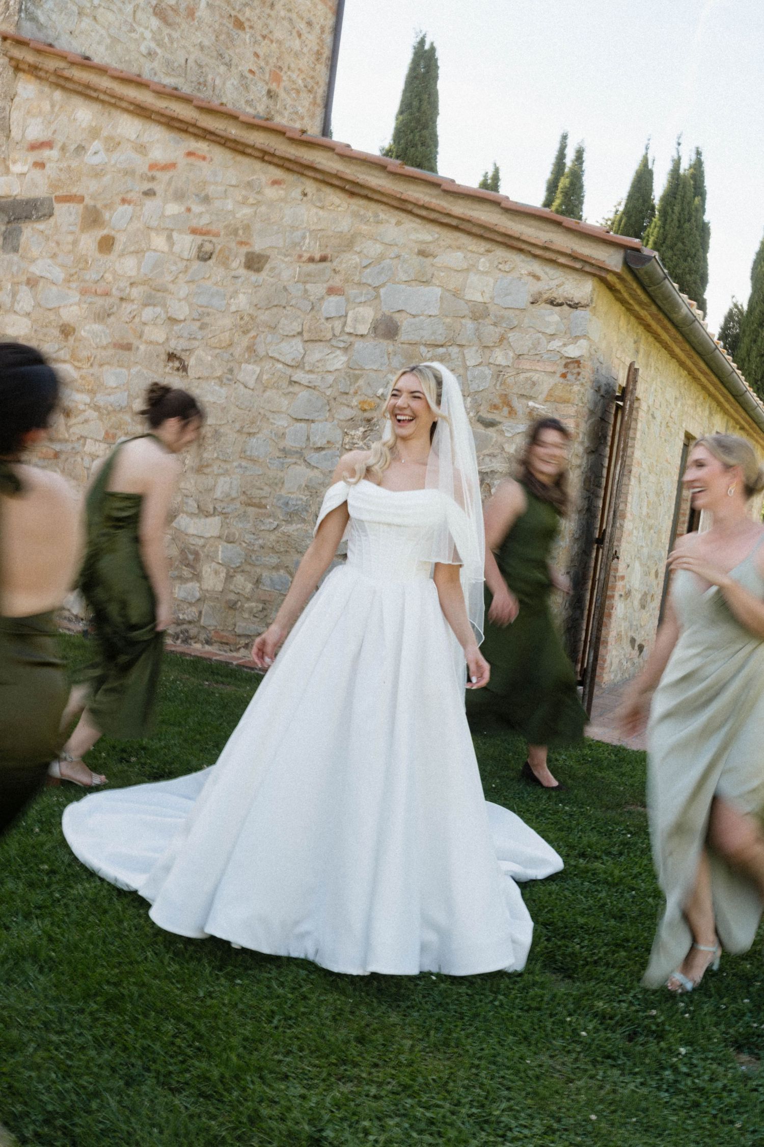 Bride in white off-shoulder gown laughing with bridesmaids in olive green dresses outside a Tuscan stone villa.