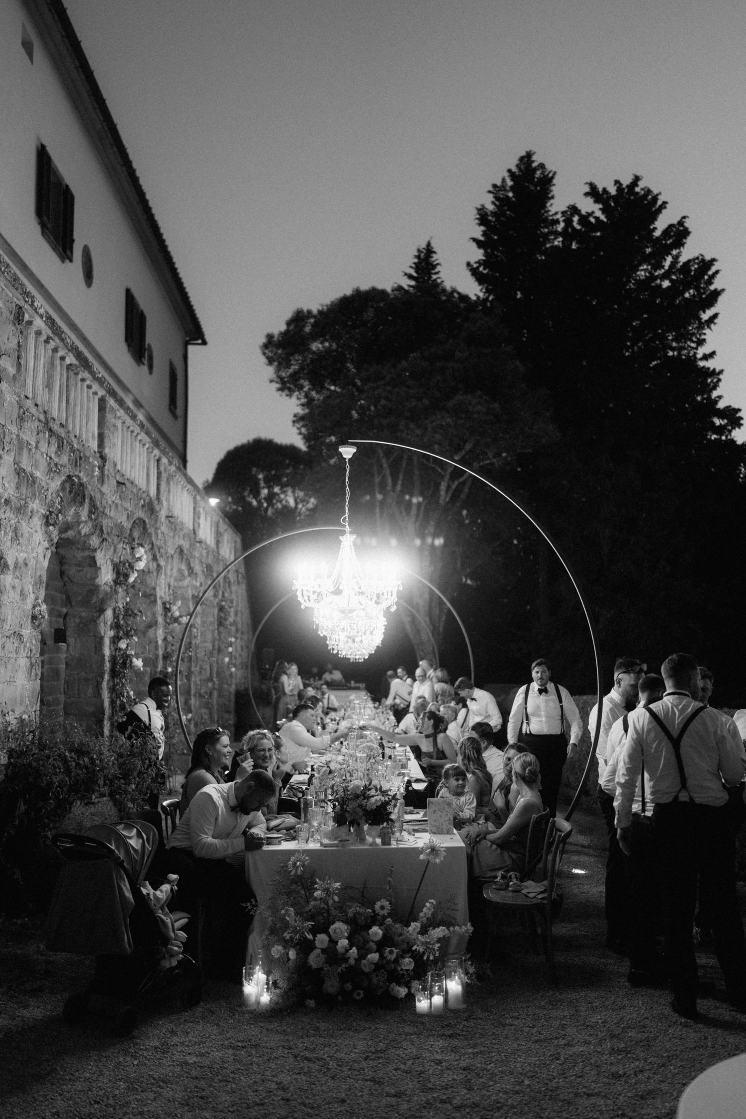 Black and white photo of an outdoor wedding reception dinner under a chandelier at dusk.