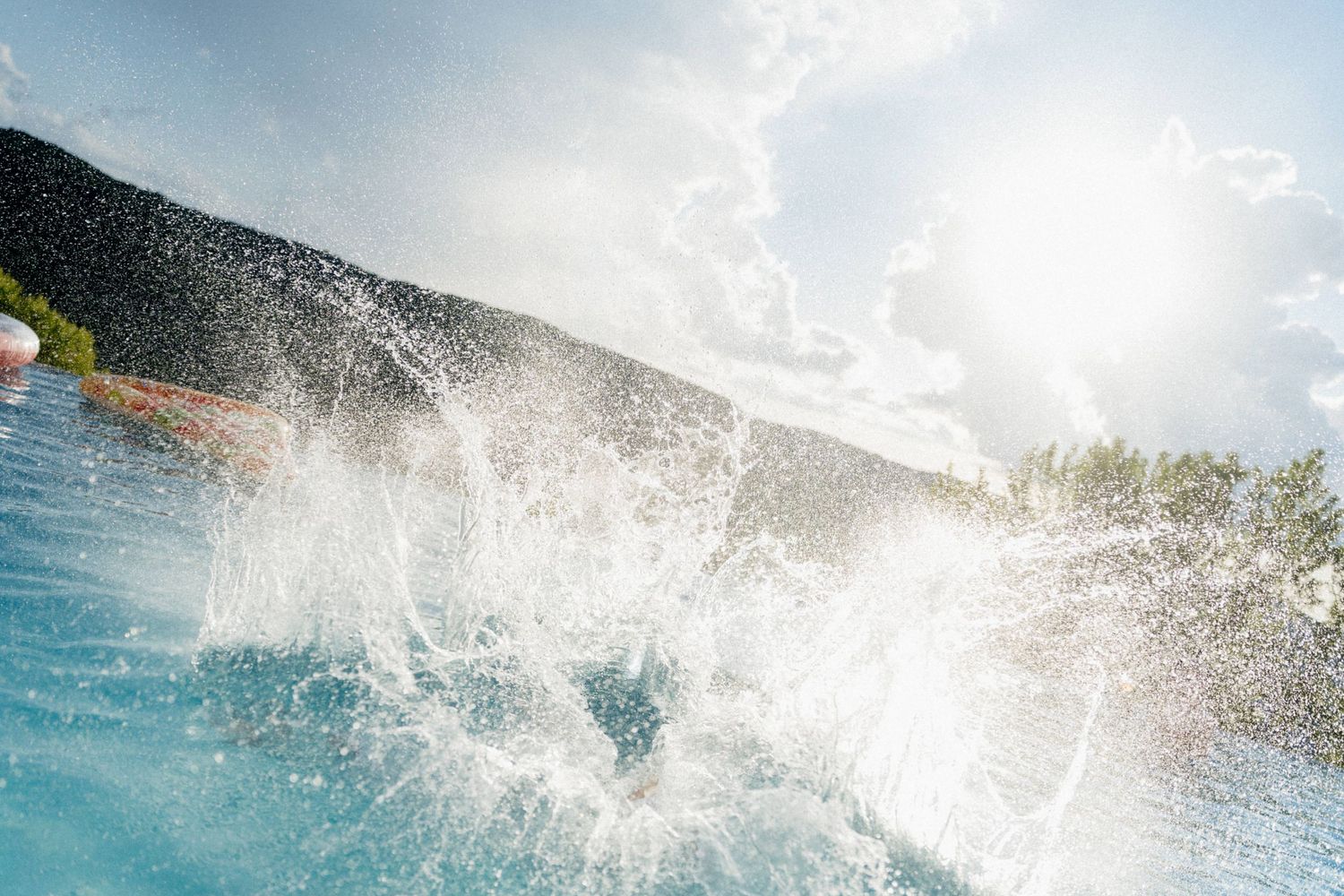 Dynamic action shot of a person splashing into a bright blue outdoor pool with mountain backdrop.