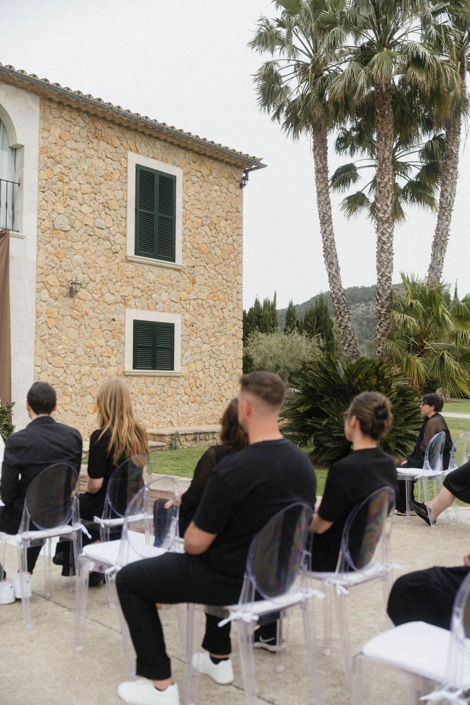 Guests in black attire seated on ghost chairs at an outdoor wedding ceremony beside a stone villa with palm trees.