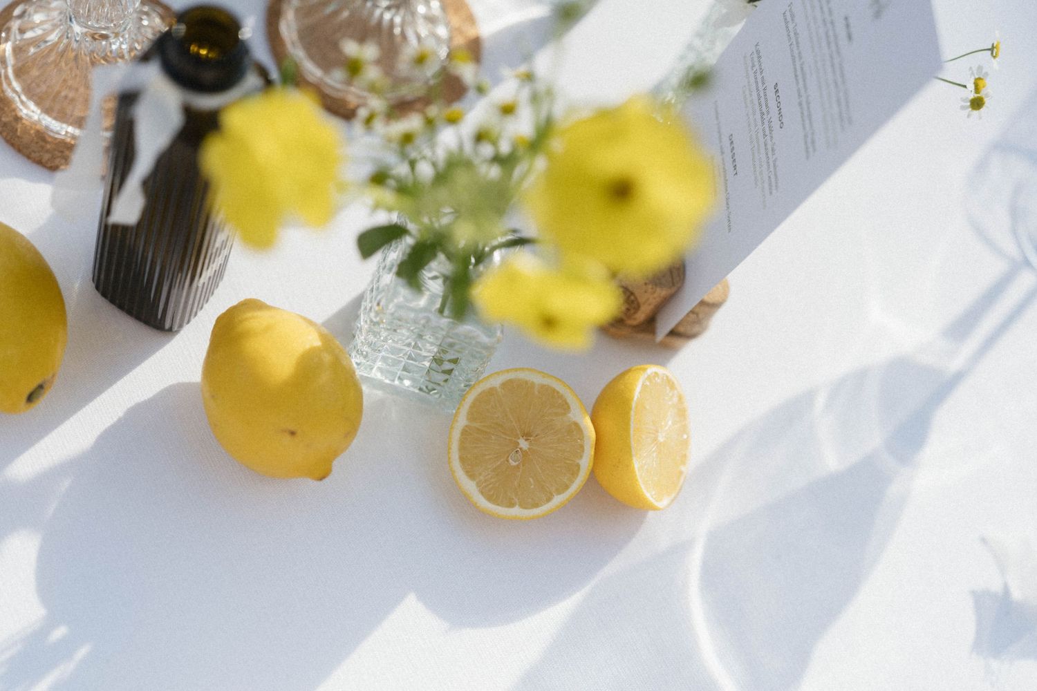 Fresh yellow lemons, one halved, displayed on a white surface with small yellow flowers in a glass vase.
