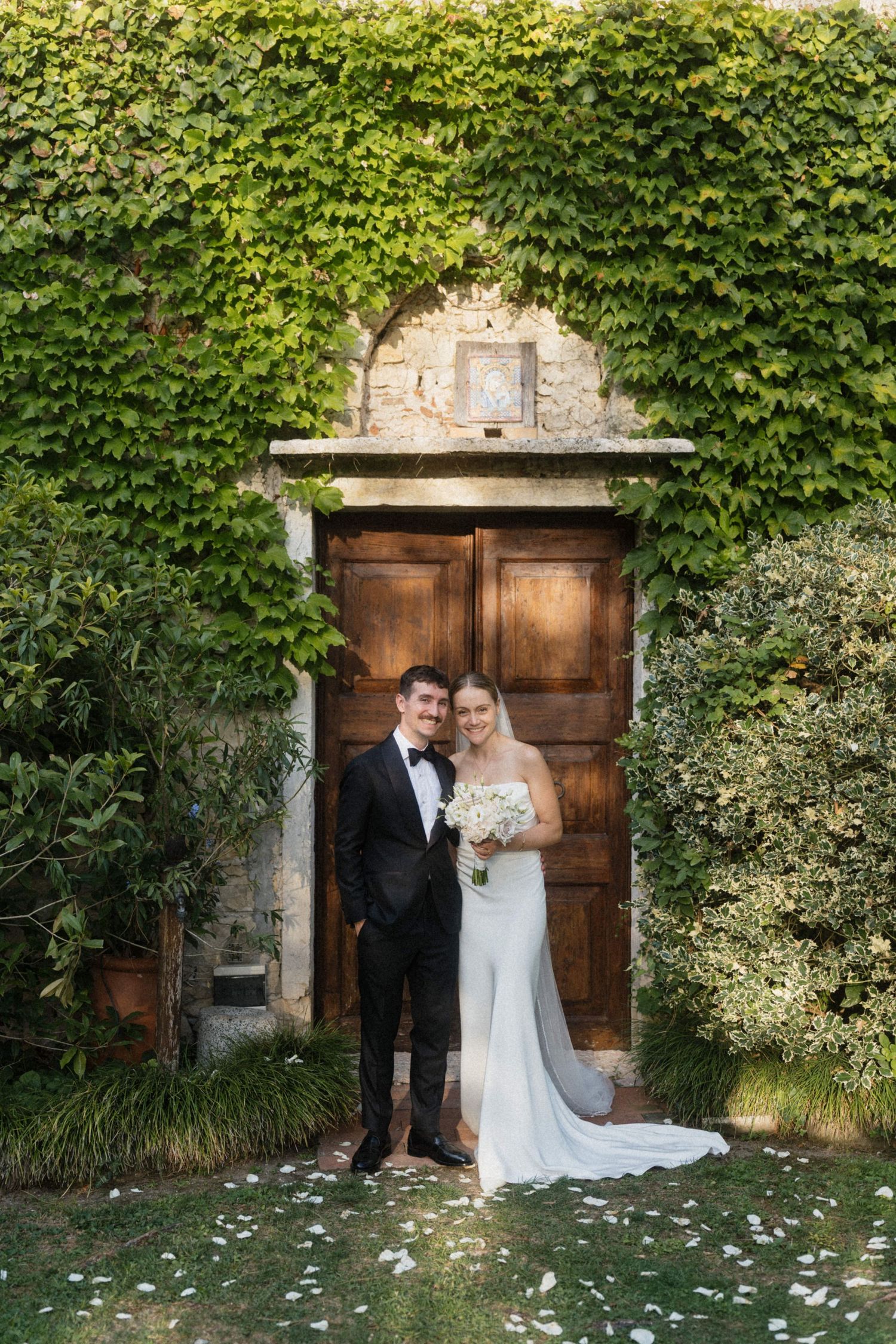 Smiling bride and groom pose before an ivy-covered stone wall and wooden arched door at their wedding.