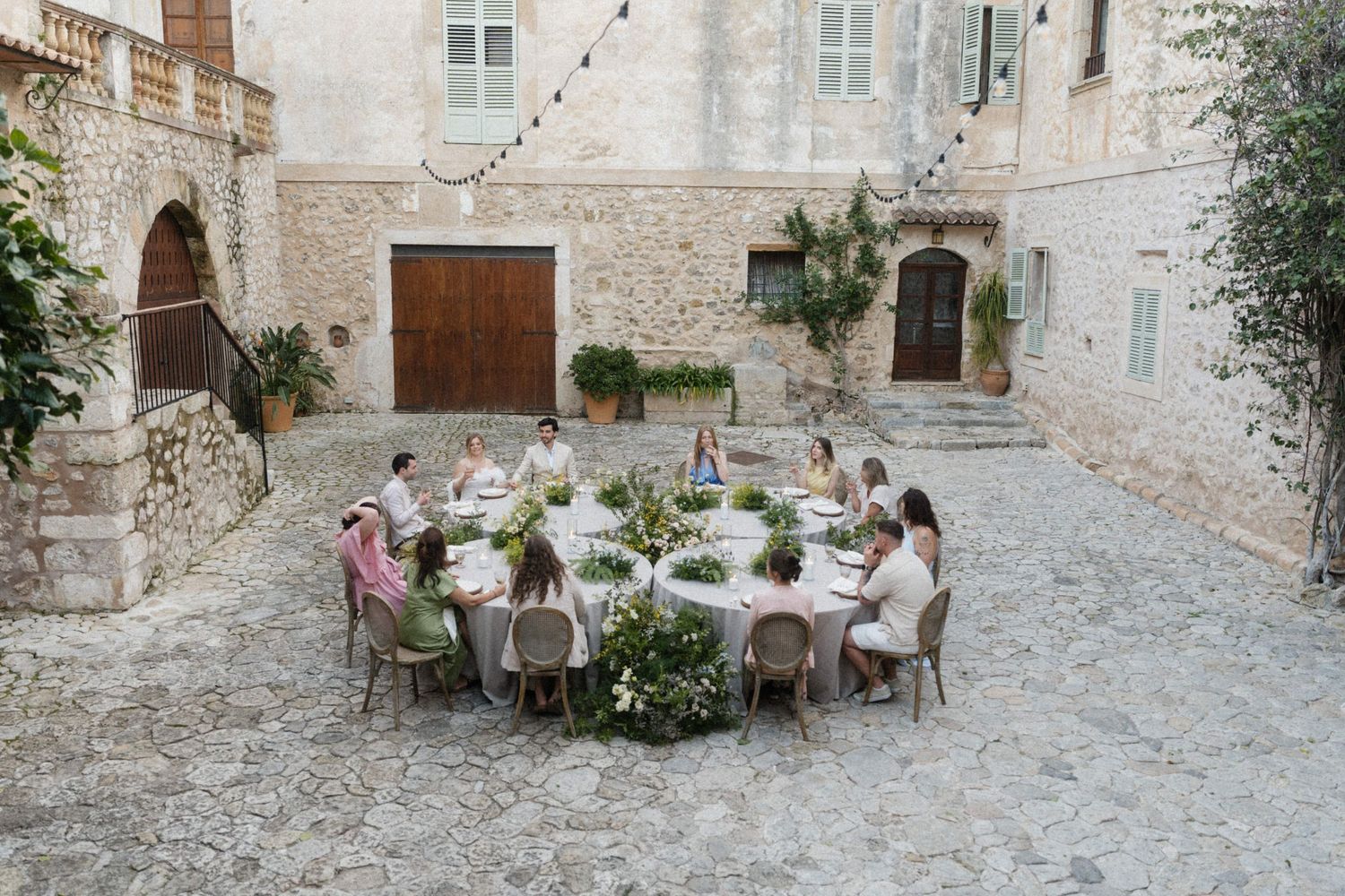 Guests dining at a round table in a rustic stone courtyard adorned with lush greenery during an outdoor wedding.
