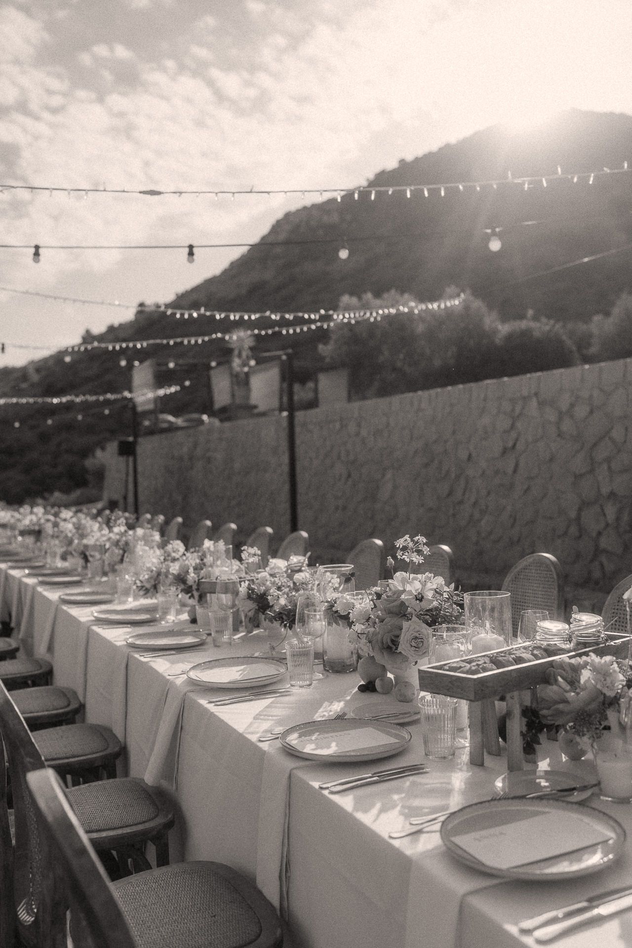 Black and white photo of an outdoor wedding reception table set with flowers, string lights, and hillside backdrop.