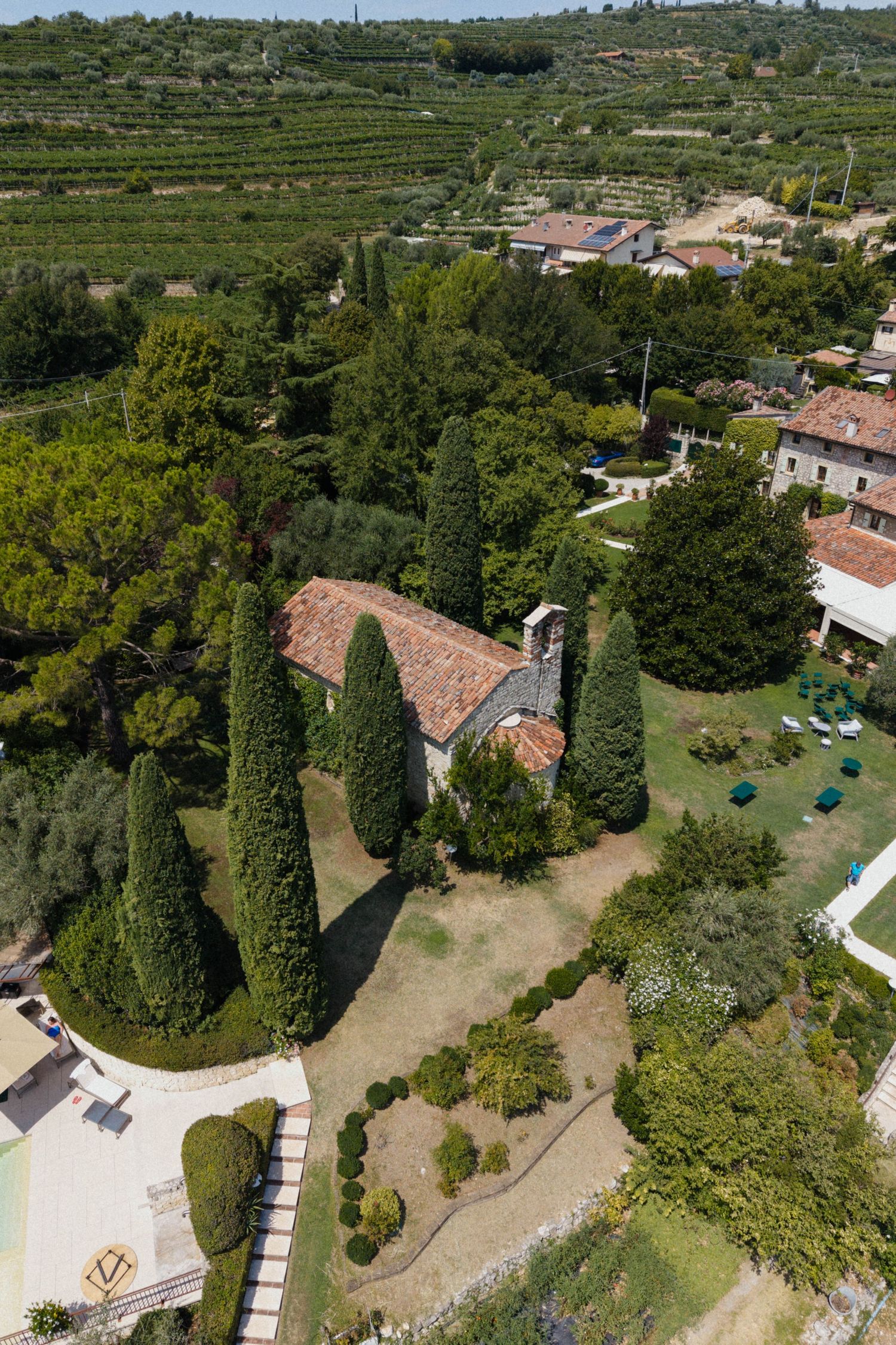 Aerial view of a historic stone chapel surrounded by cypress trees and lush gardens in the Italian countryside.