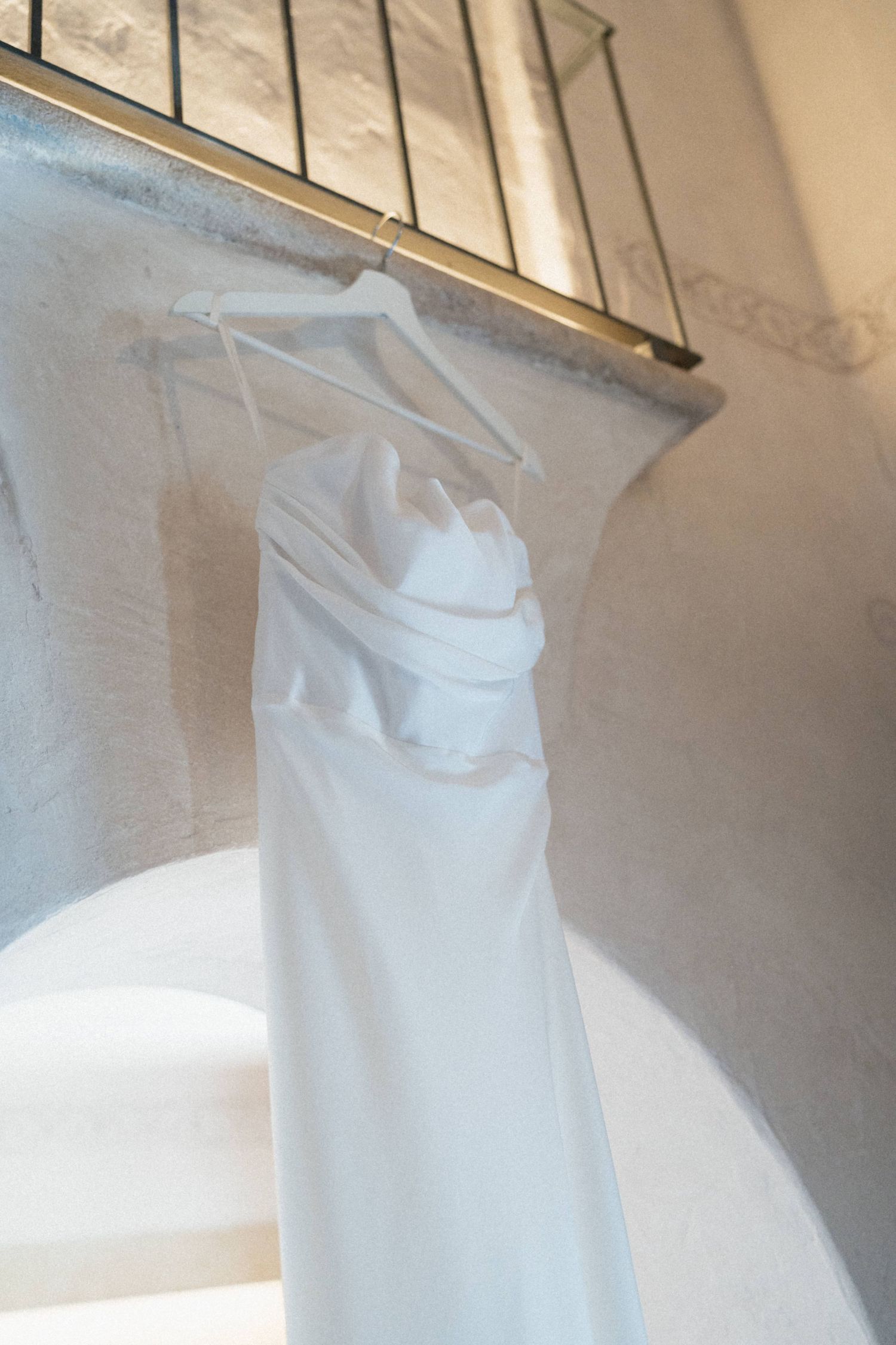 Elegant white wedding dress hanging on a hanger against a sunlit stone arch wall.