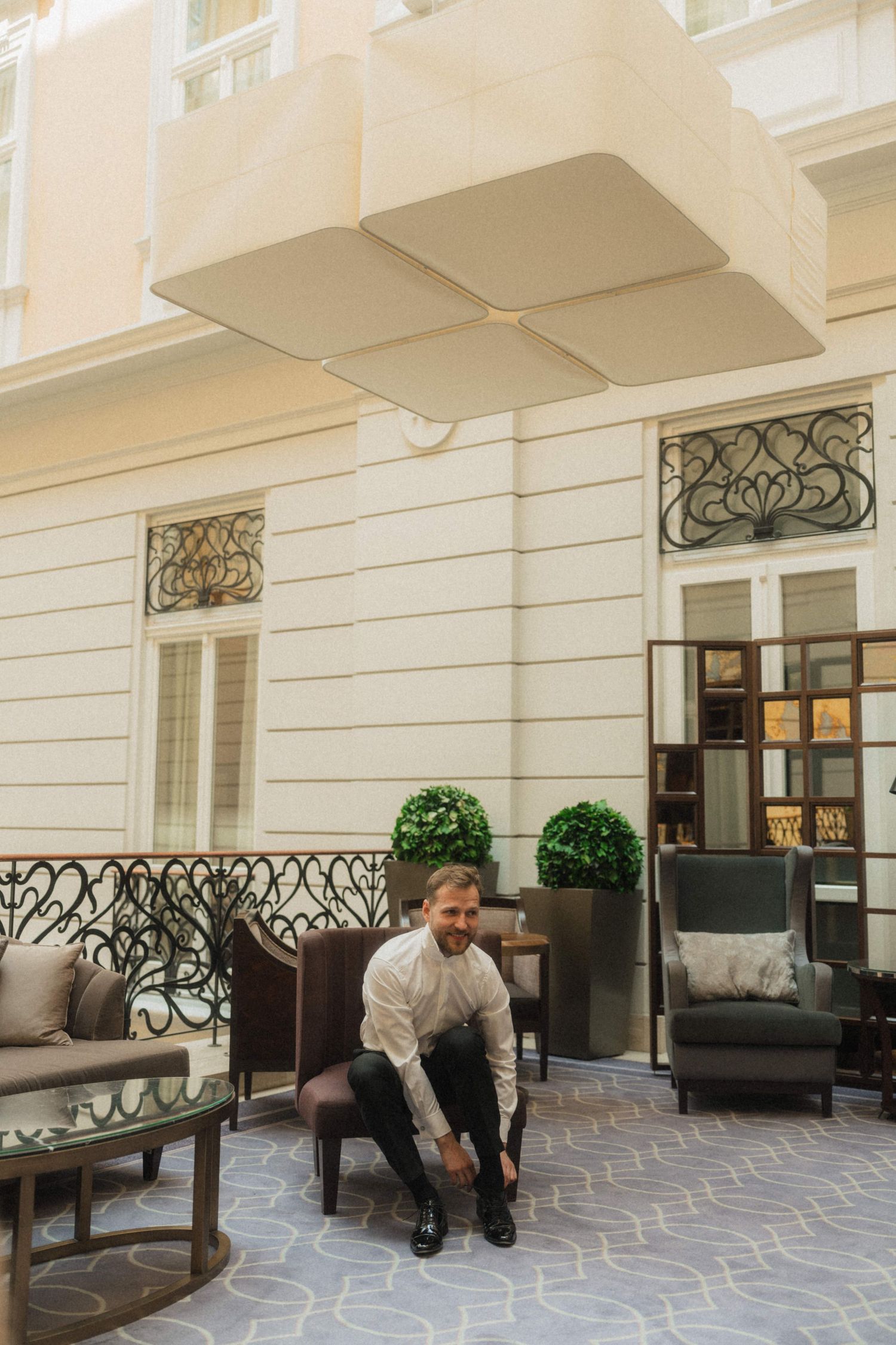 Groom tying his shoes in an elegant hotel courtyard with ornate iron railings and lush greenery.