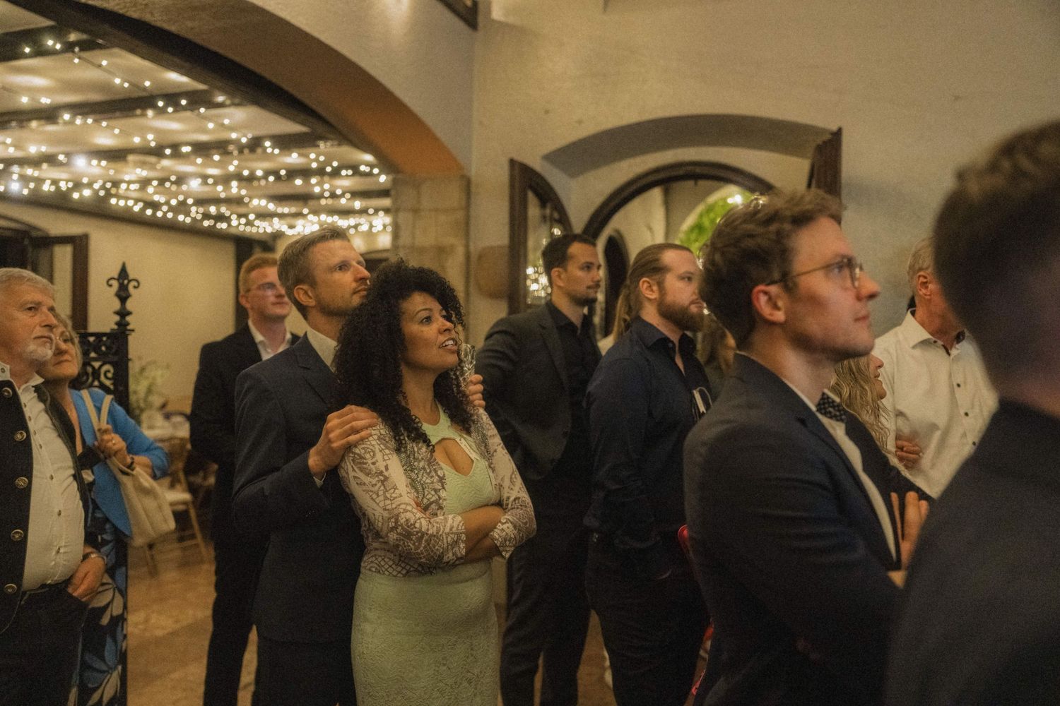 Wedding guests gathered indoors under string lights in rustic arched venue during reception.