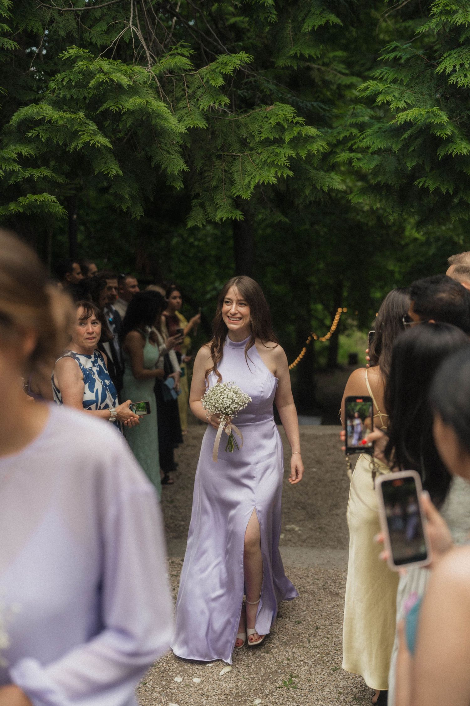 Smiling bridesmaid in lavender satin gown walks down outdoor aisle holding white bouquet among wedding guests.