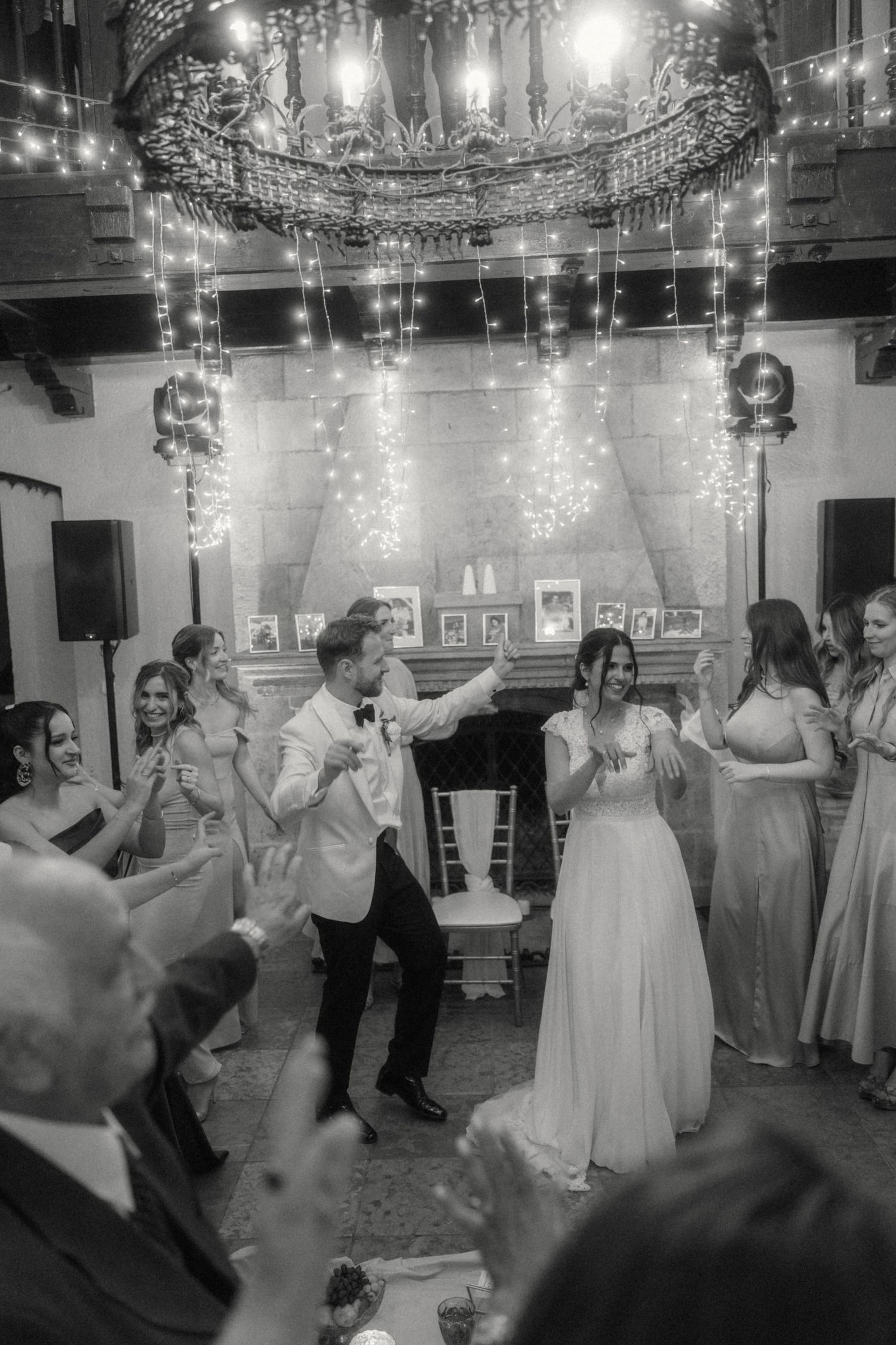 Bride and groom dancing joyfully at wedding reception in black and white photo with string lights.