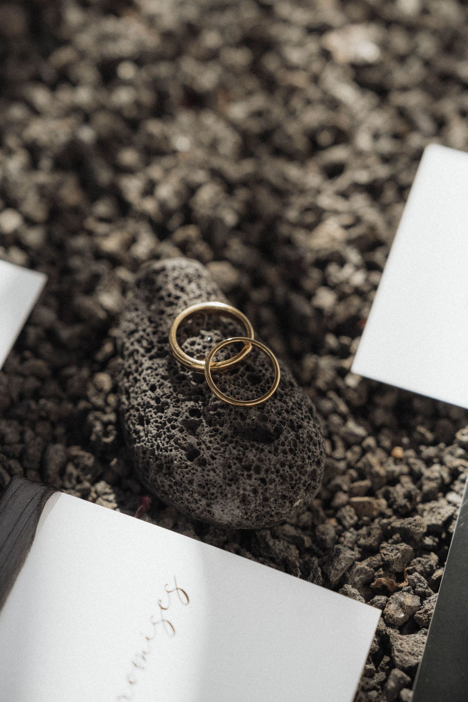 Two gold wedding rings resting on a dark volcanic rock surrounded by gravel near an open wedding vow booklet.