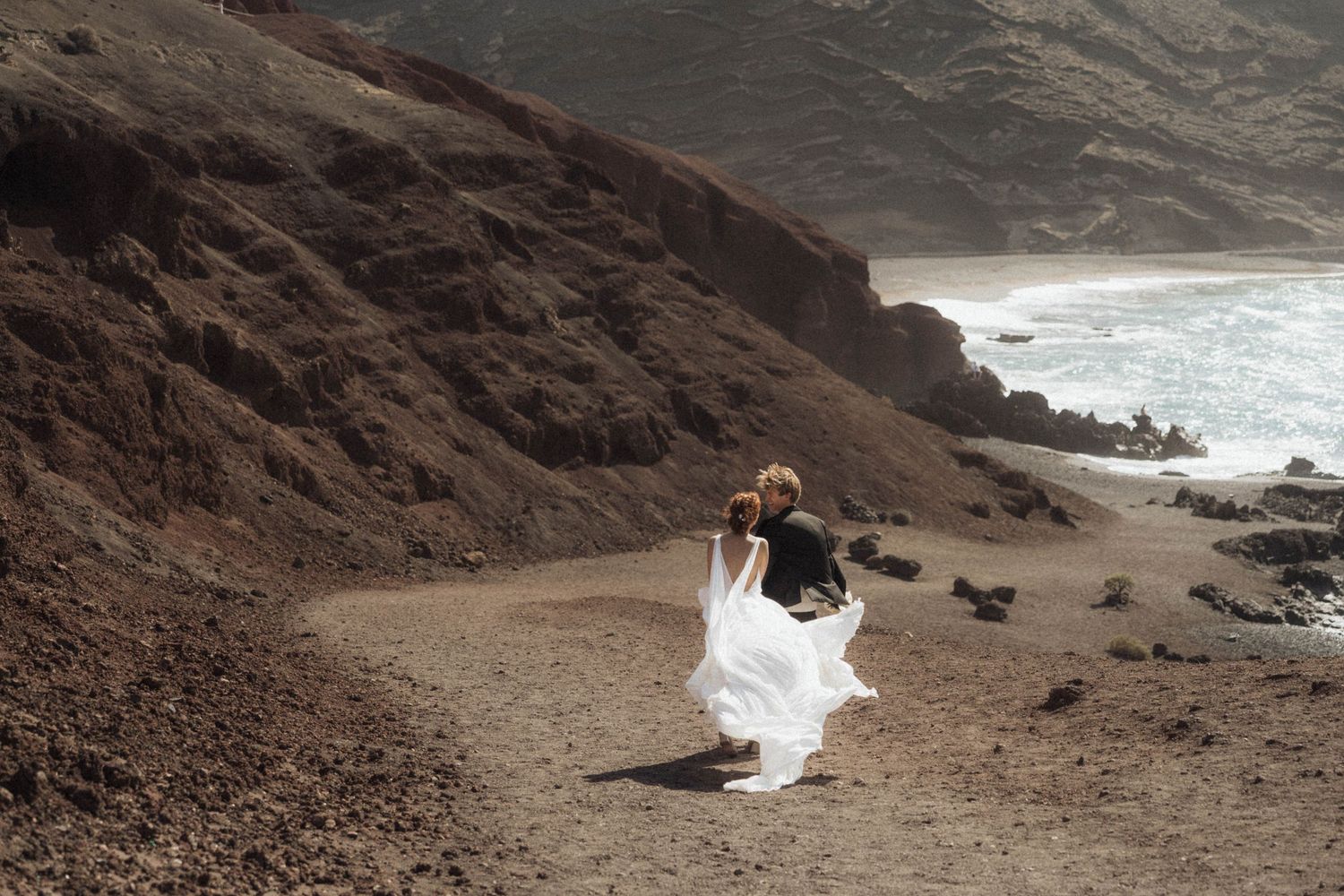 Bride and groom embracing on a rugged coastal cliff path with ocean waves below.