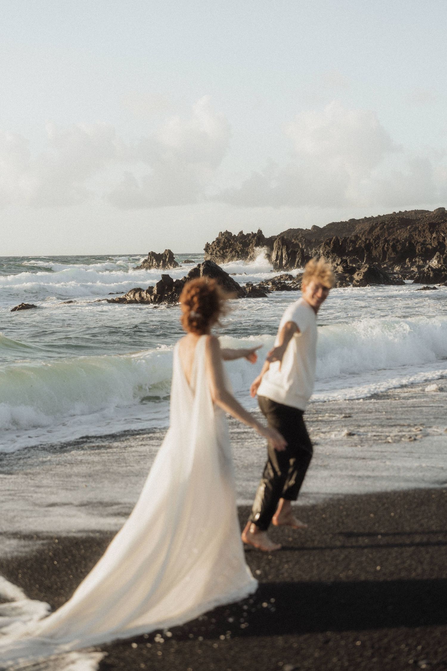 Newlyweds hold hands and walk along a black sand beach with crashing ocean waves behind them.