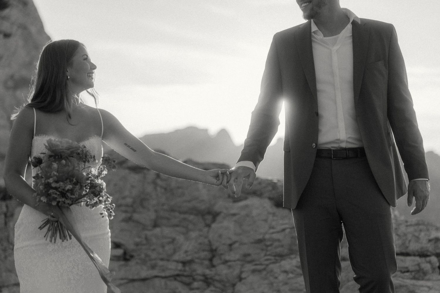 Black and white photo of a bride and groom holding hands walking outdoors with mountains in the background.