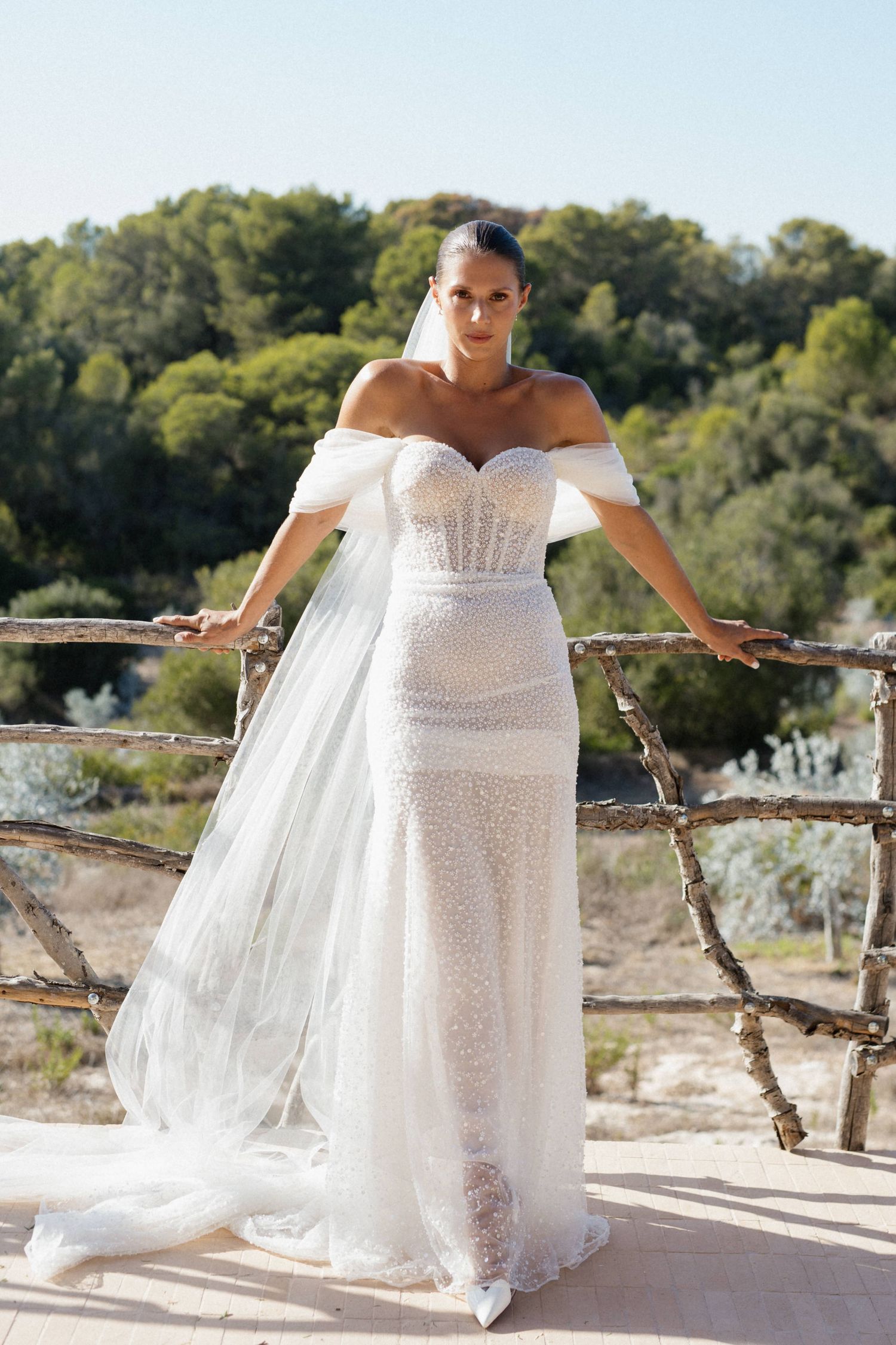 Bride in an off-the-shoulder beaded wedding gown and long veil leaning on a rustic wooden railing with a green hillside behind.
