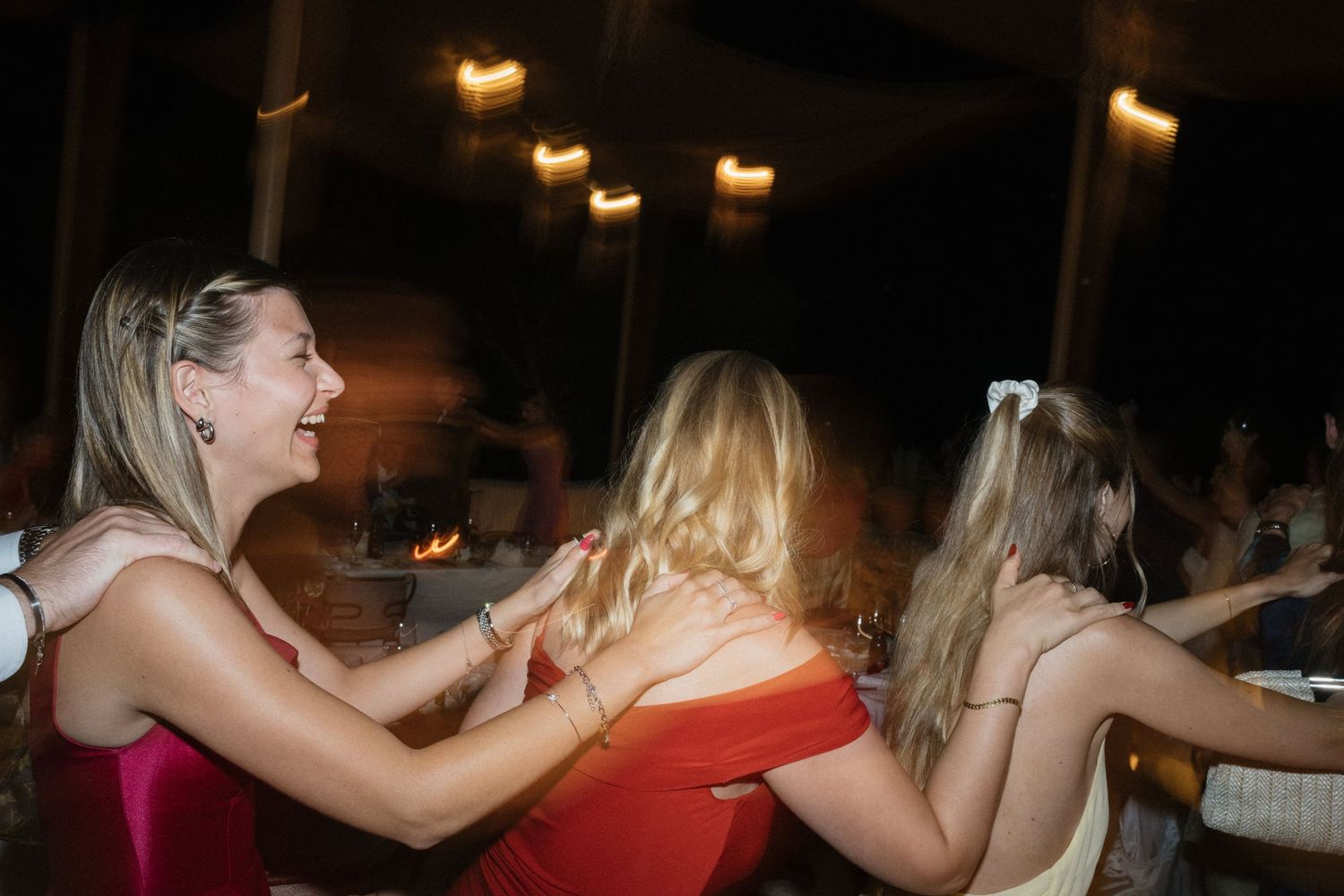 Smiling wedding guests in red and cream dresses link arms and dance together at an evening reception.