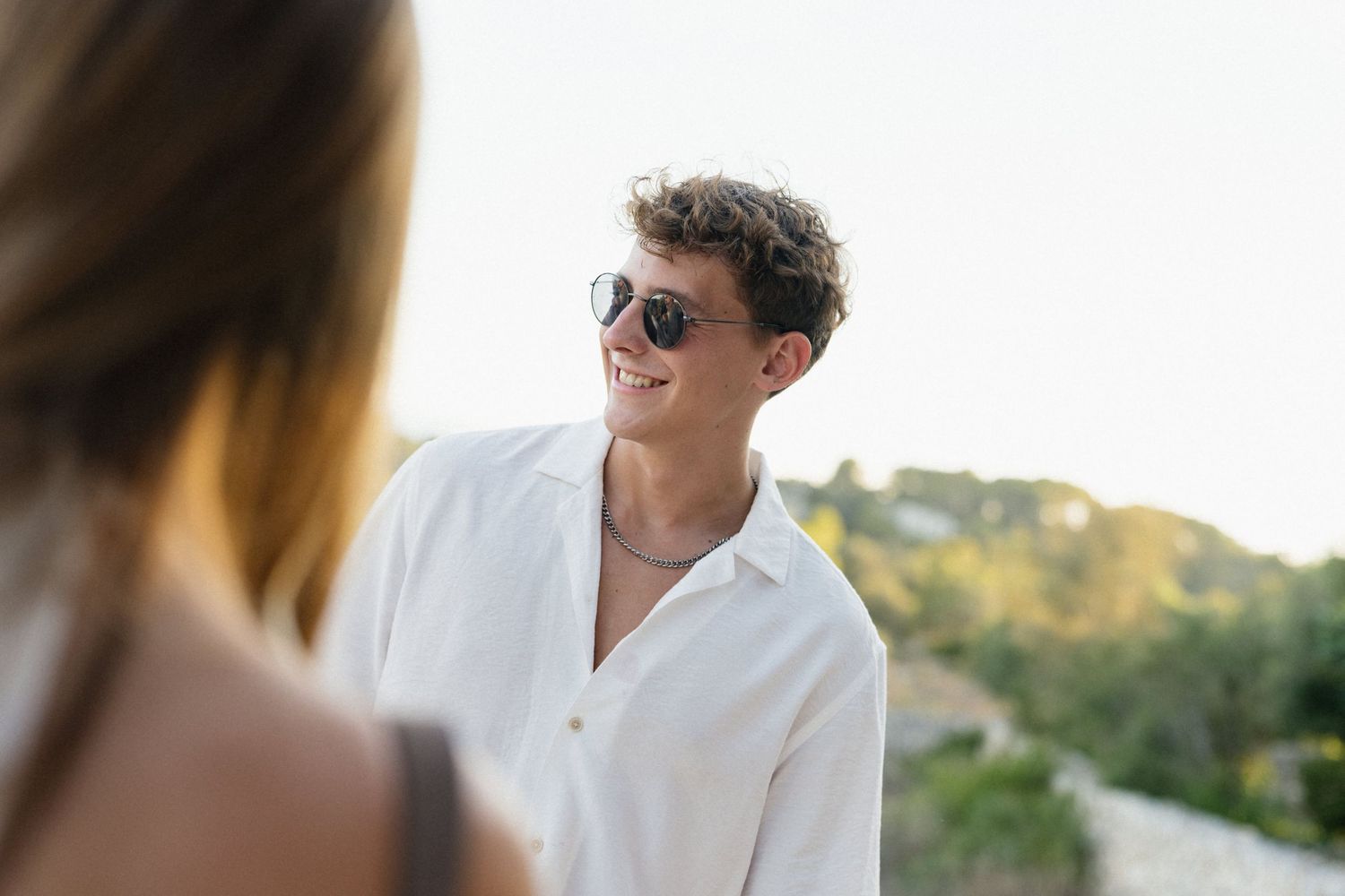 Smiling young man in sunglasses and a white linen shirt stands outdoors on a sunny day with a blurred woman in the foreground.