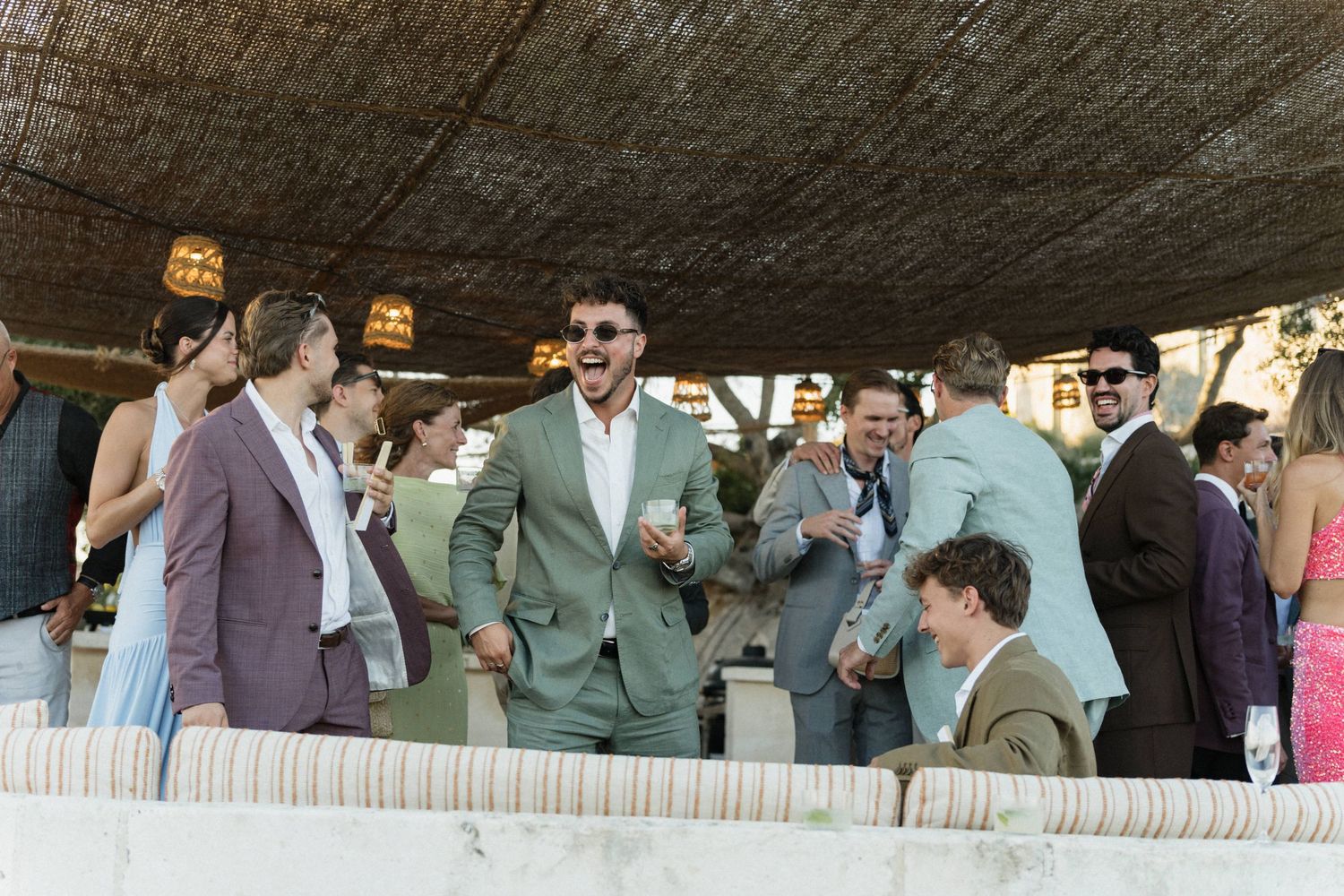 Smiling guests in suits and sunglasses laugh and mingle with drinks during an outdoor wedding reception under a shaded canopy.