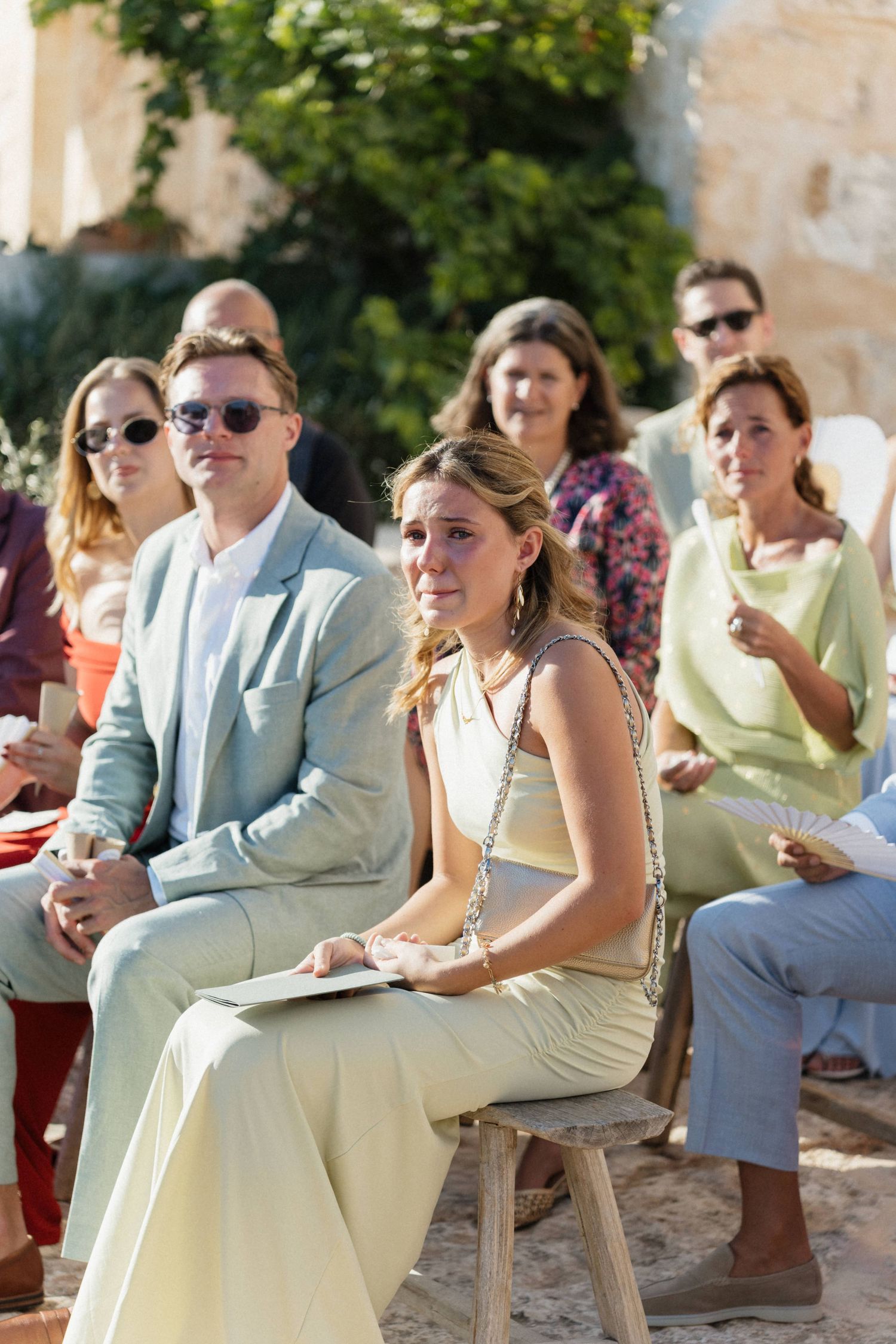 Wedding guests in pastel outfits sit on wooden benches, a woman in cream looking emotional during an outdoor ceremony.