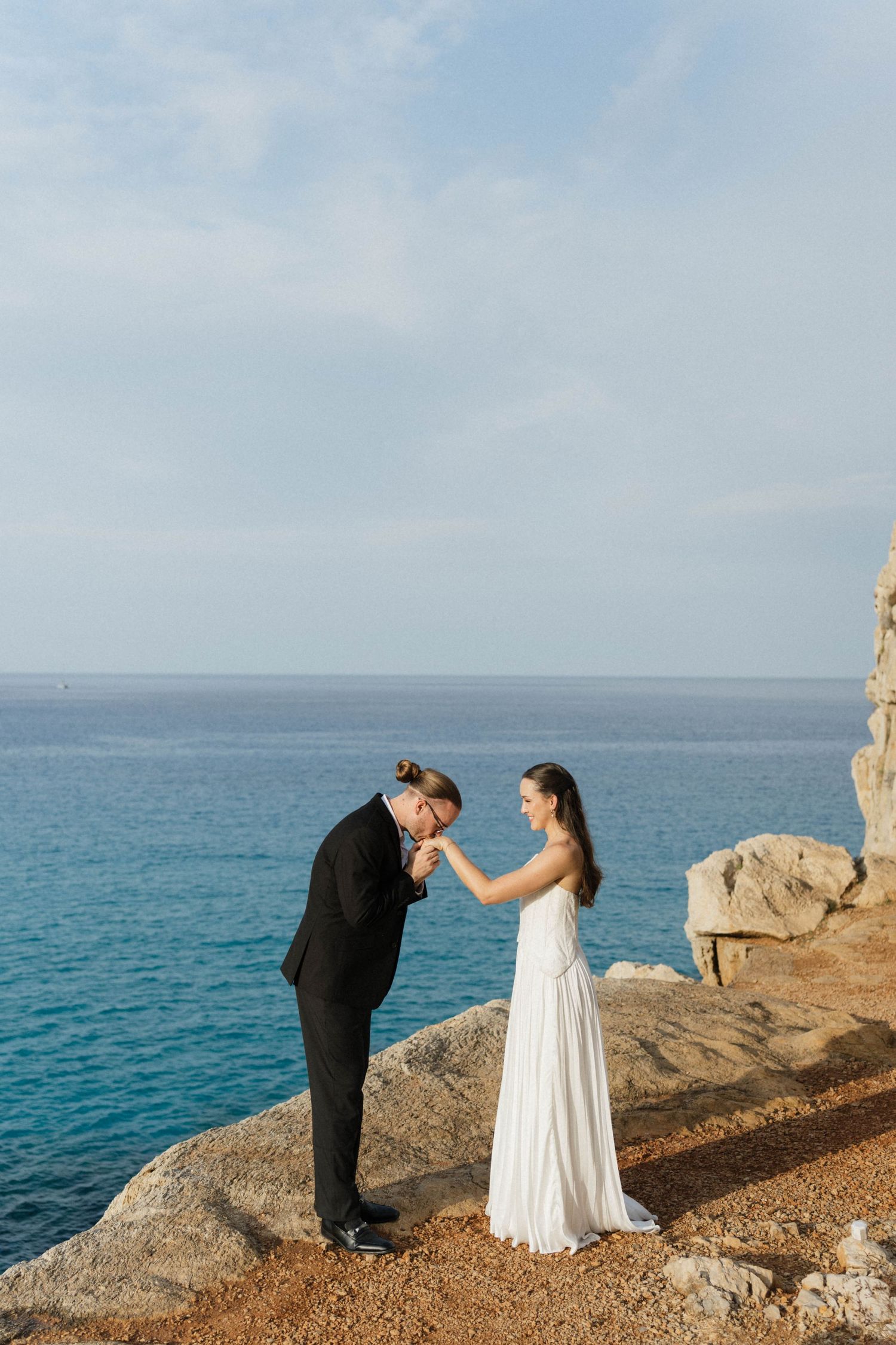Groom bows to kiss his bride's hand as she stands in a white dress on a coastal cliff overlooking the ocean.
