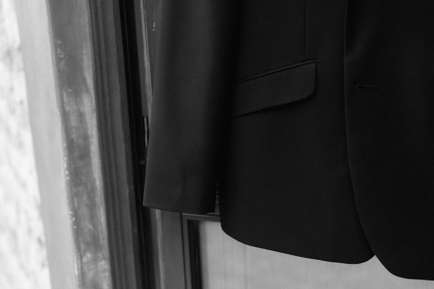 Black suit jacket with a chest pocket hanging on a hanger beside a window in a black and white photo.