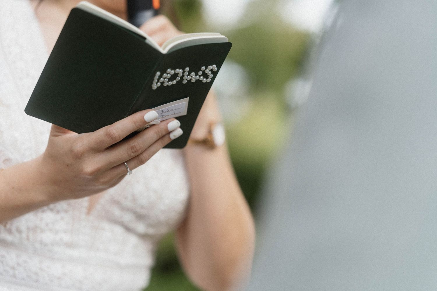 Close-up of a bride in white lace dress holding a black vow book during an outdoor wedding ceremony.