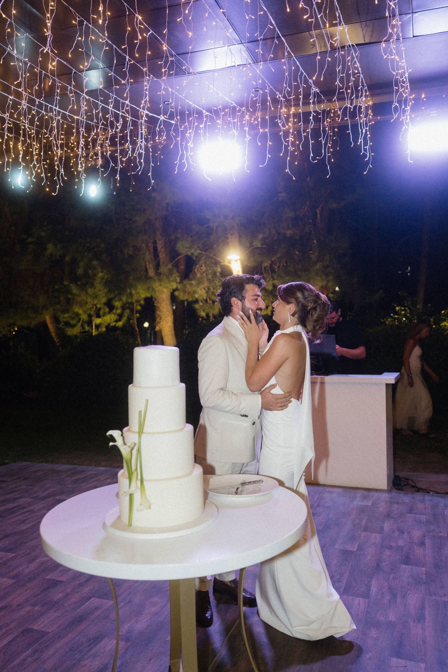 Bride and groom share a kiss beside a four-tier white wedding cake under sparkling fairy lights at night.