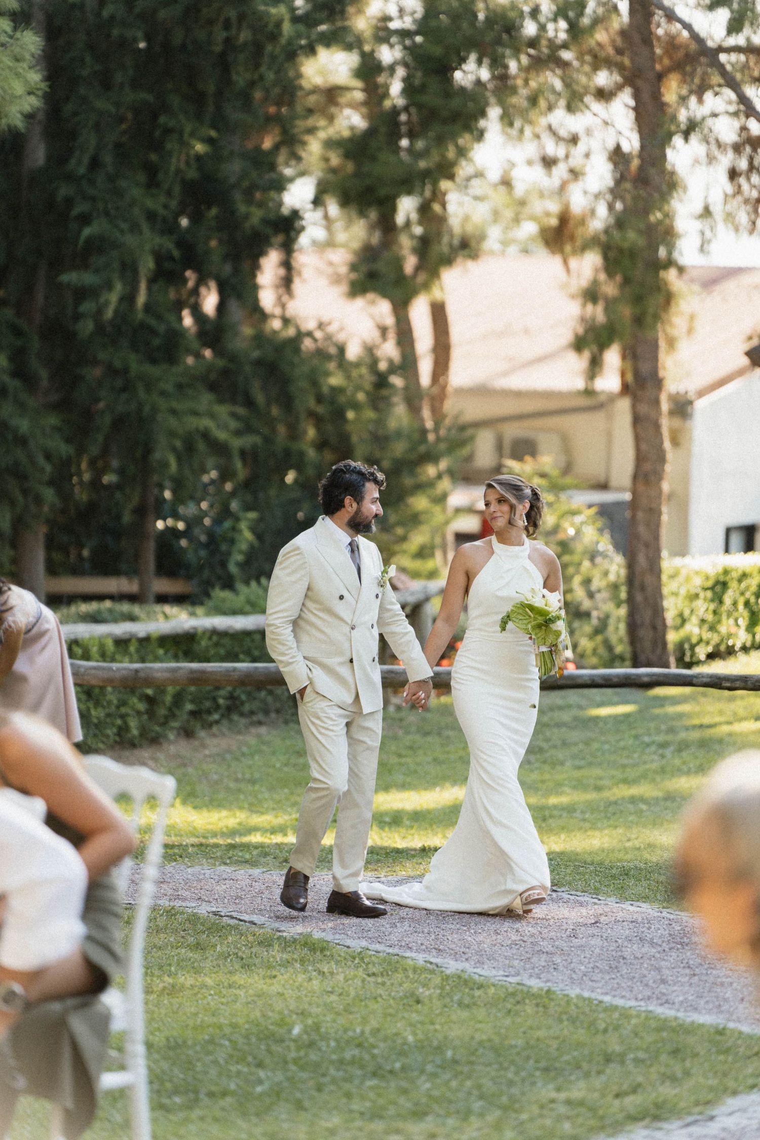 Bride and groom walk hand-in-hand down outdoor aisle, smiling at each other, surrounded by lush green trees.