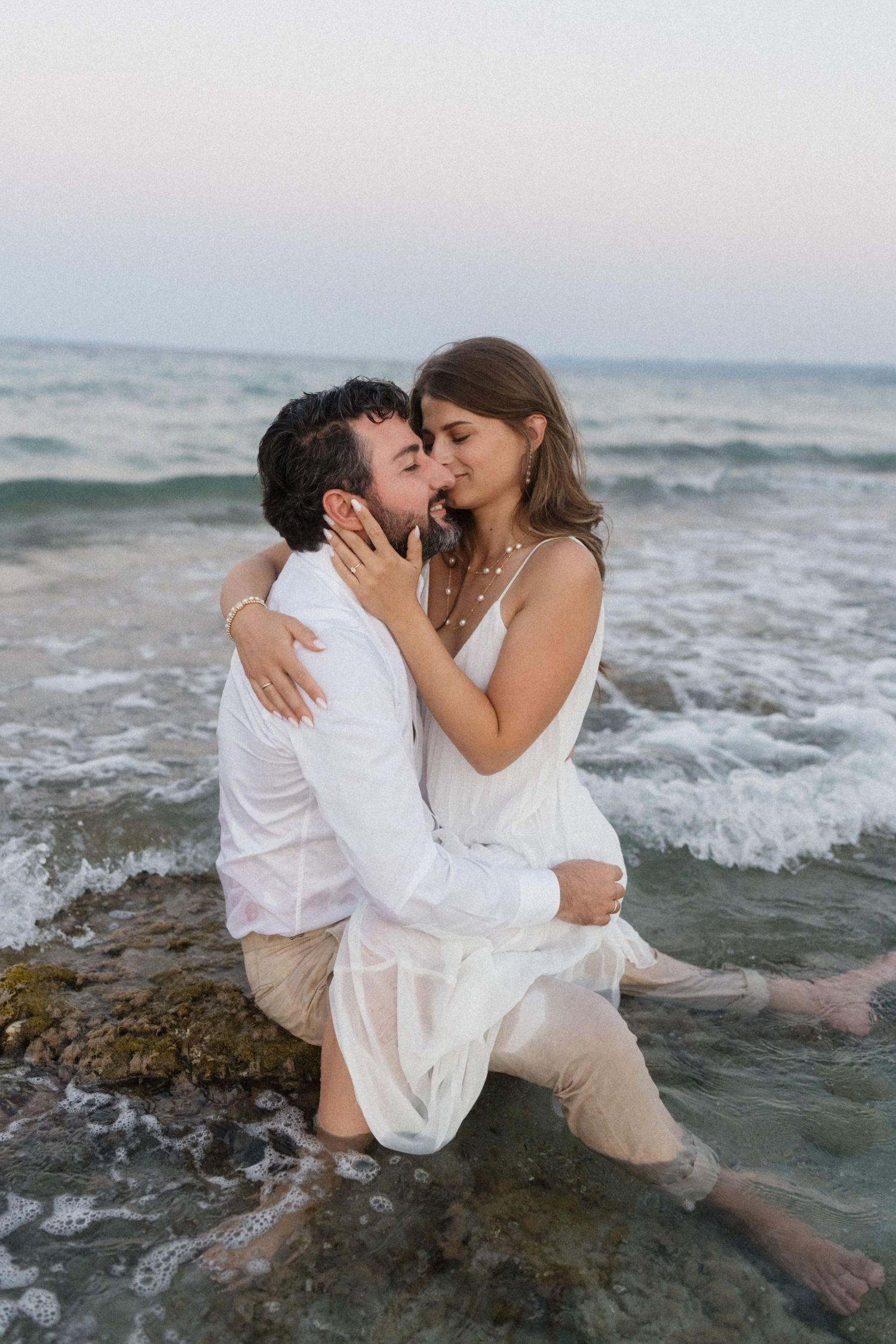 Couple sharing a romantic kiss while sitting in ocean waves on a rocky beach at sunset.