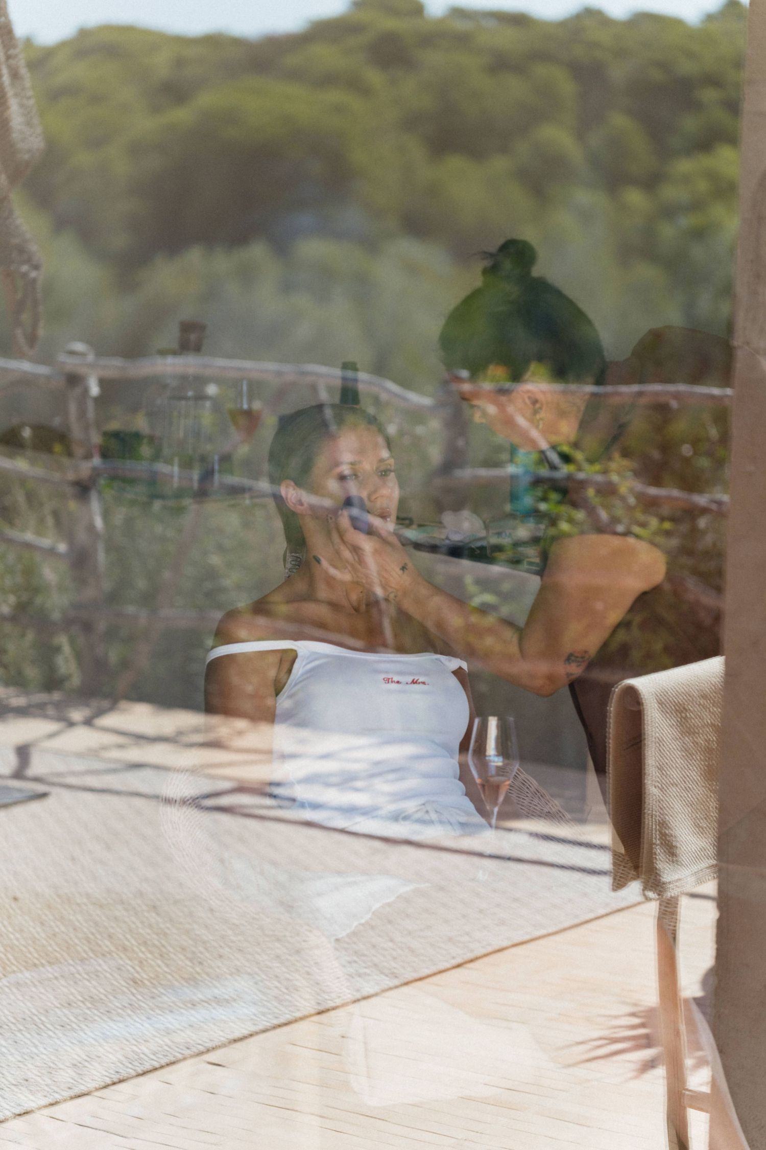 Woman in a white tank top receives a makeup touch-up on a sunlit wooden terrace, seen through glass with forest reflections.