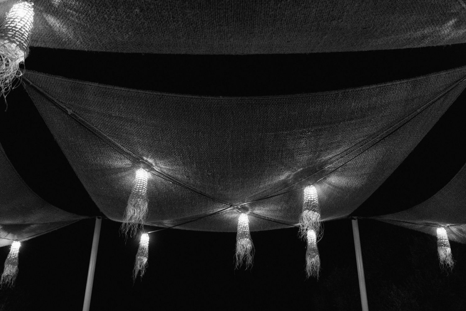 Black and white photo of fabric canopies with glowing tassel lanterns hanging overhead at an evening wedding reception.