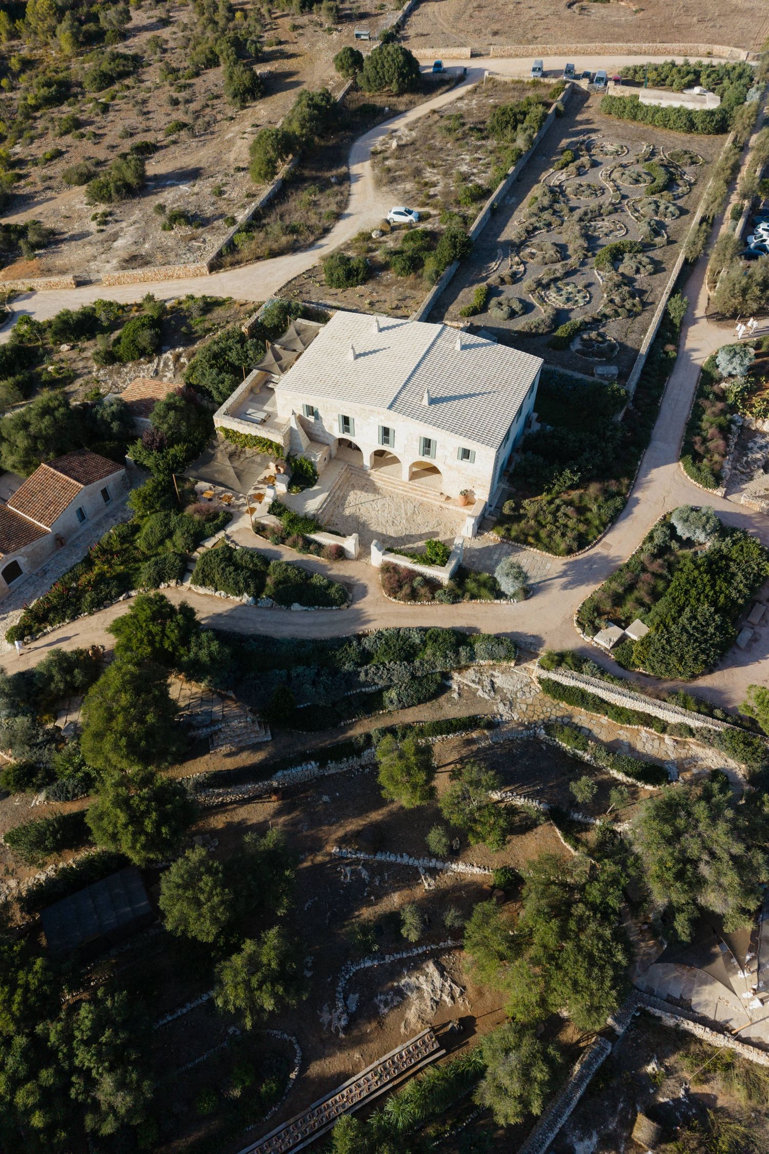 Aerial view of a white stone villa with arched loggia, surrounded by terraced gardens, olive trees, dry stone walls and winding paths.