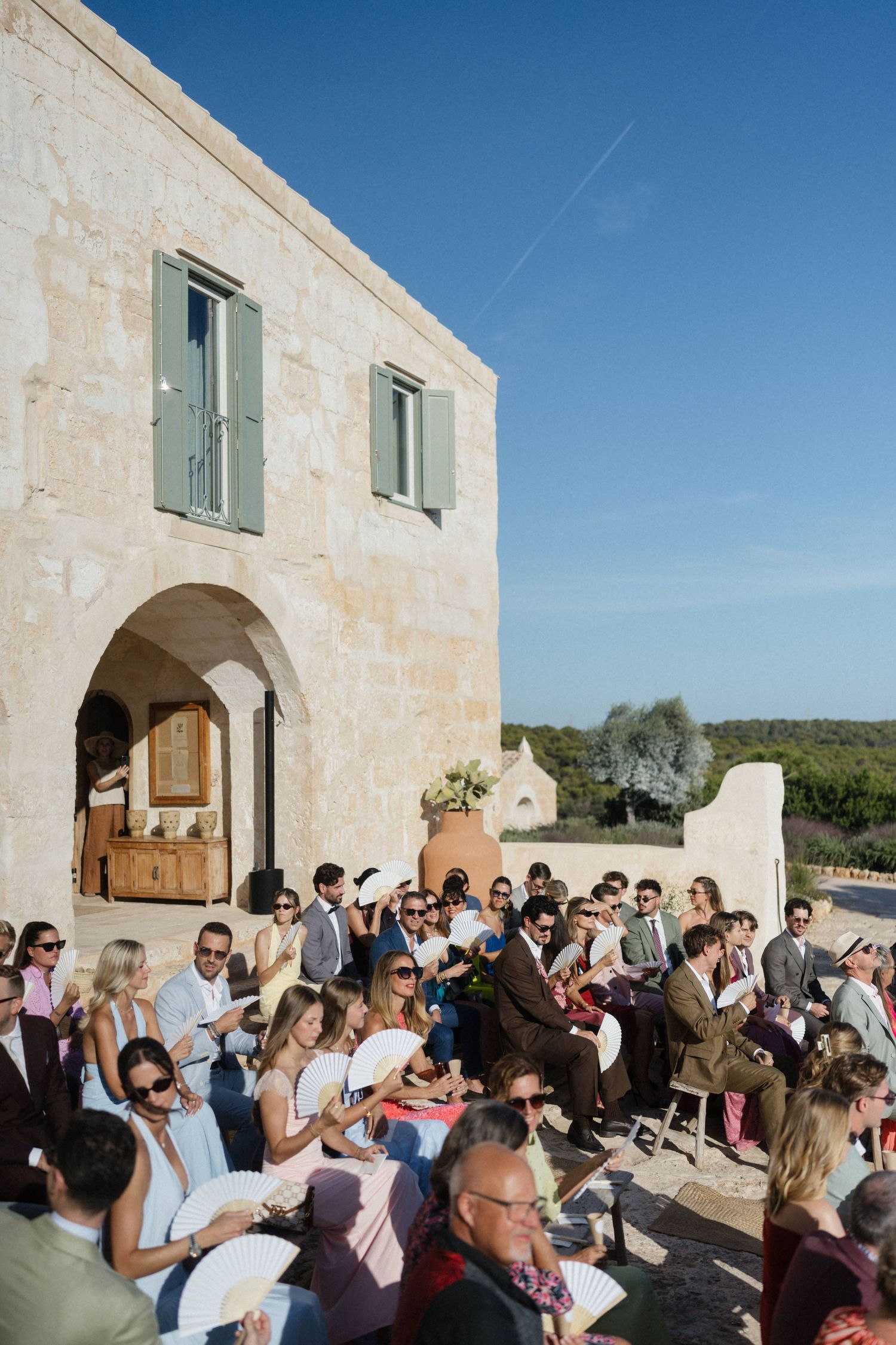 Wedding guests in summer attire hold fans and watch an outdoor ceremony beside a rustic stone villa with green shutters.