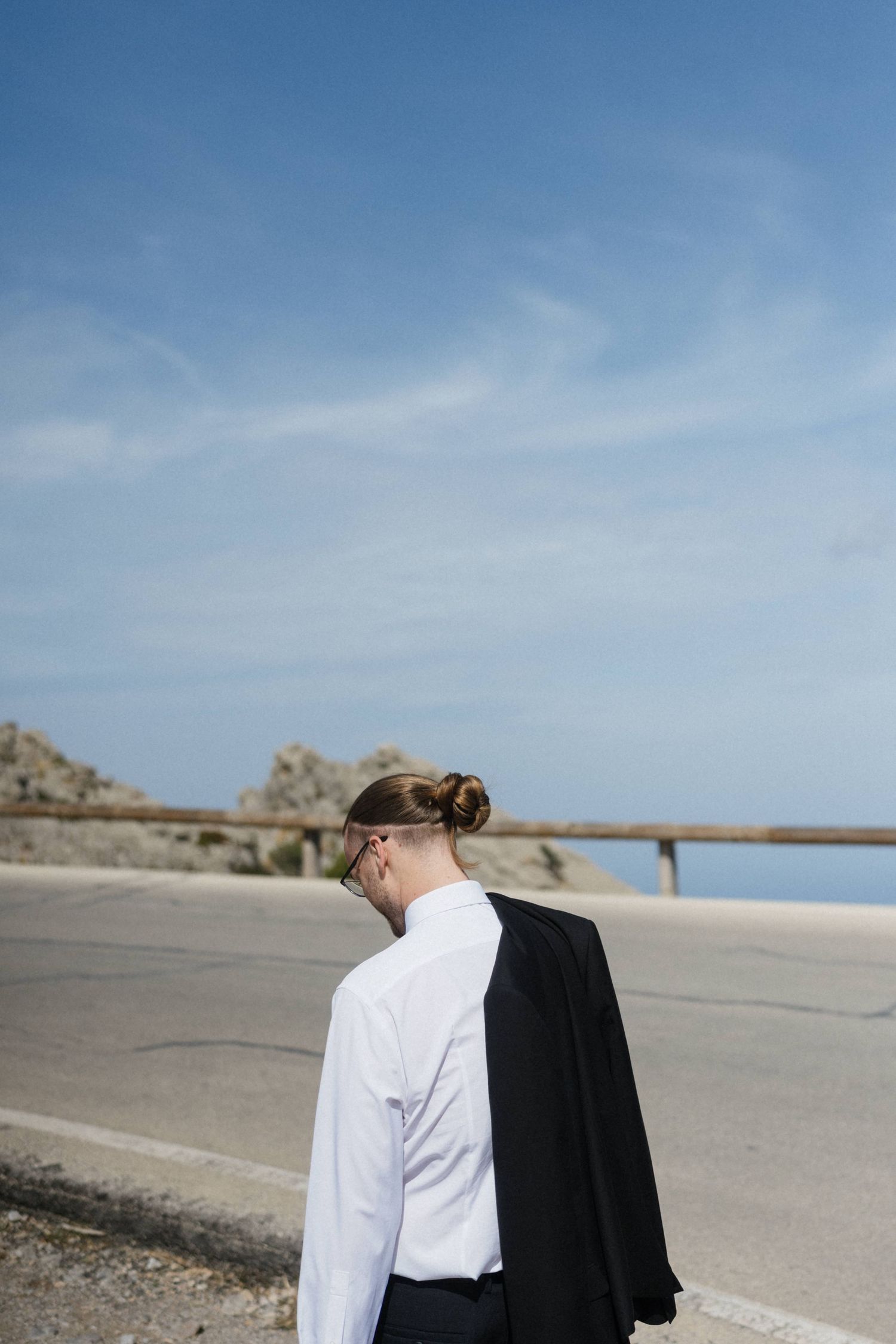 Man with a hair bun and glasses in a white shirt carries a black jacket over his shoulder on a coastal road under a blue sky.