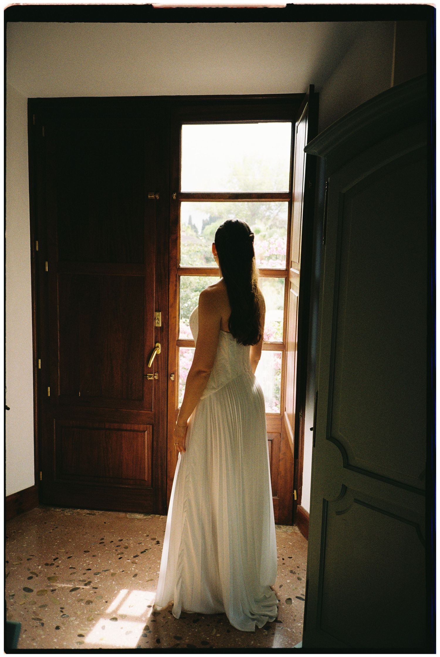 Bride in a flowing white wedding gown stands facing an open wooden door, with sunlight streaming across the speckled terrazzo floor.