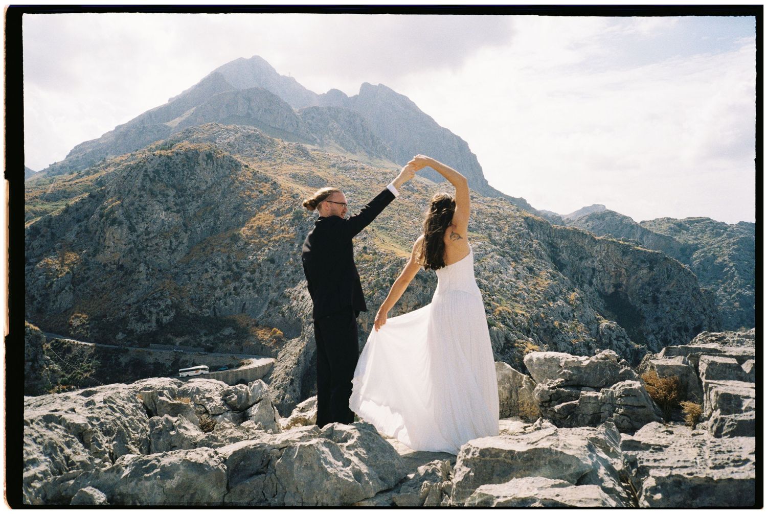 Bride in a white gown and groom in a black suit dance hand in hand on rocky mountain terrain during an outdoor elopement photo shoot.