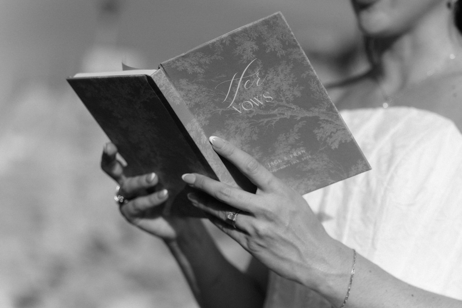 Black and white photo of a woman in a light top holding an open vow book with a decorative cover, wearing rings and a bracelet.