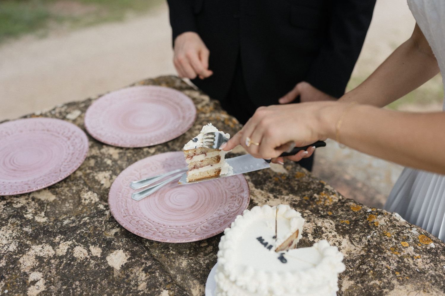 Newlyweds cutting a white frosted wedding cake with a silver knife on a rustic stone table beside pink plates outdoors.