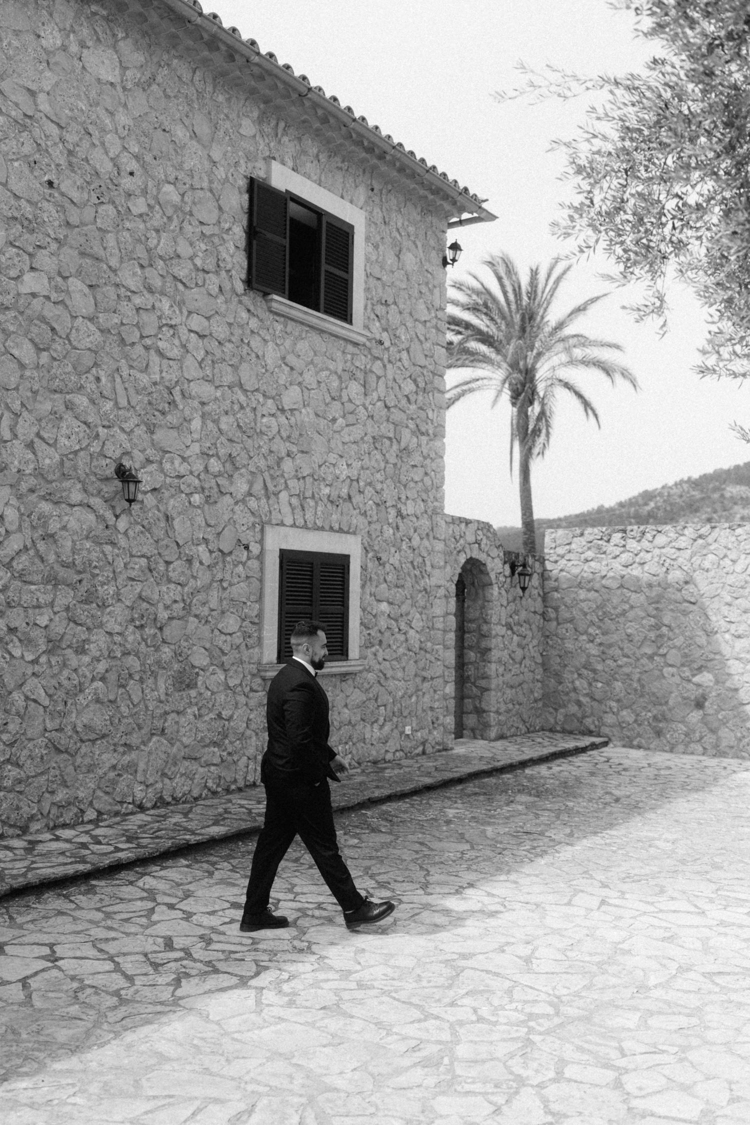 Black and white photo of a suited man walking past a stone building with palm tree in Mediterranean setting.