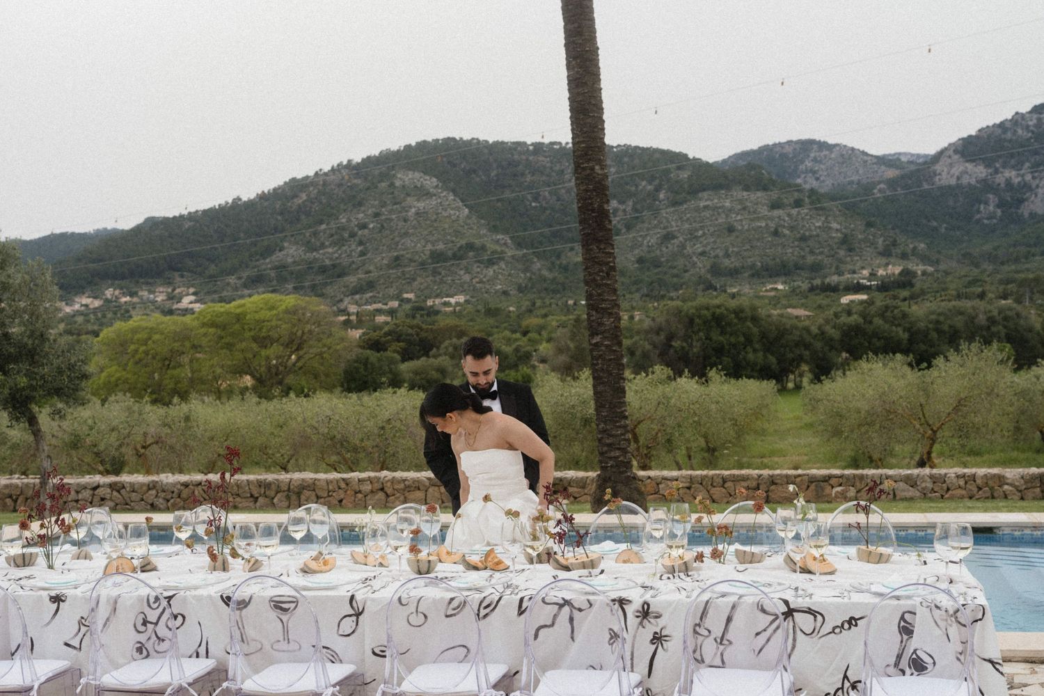 Bride and groom embrace at outdoor wedding reception table with mountain backdrop and pool view.