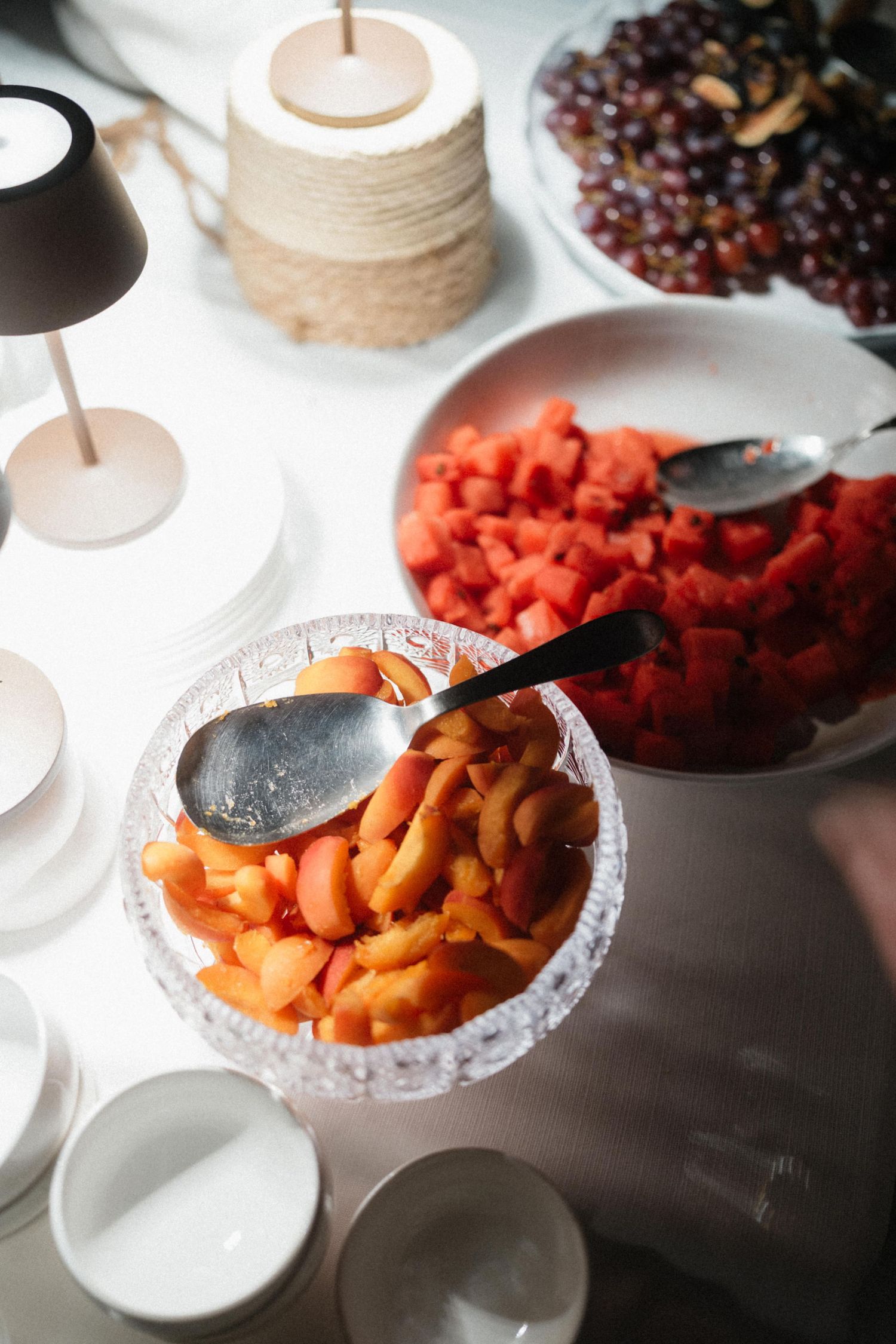 Crystal bowl with sliced peaches and melon on a white table with watermelon and grapes at a catered event.