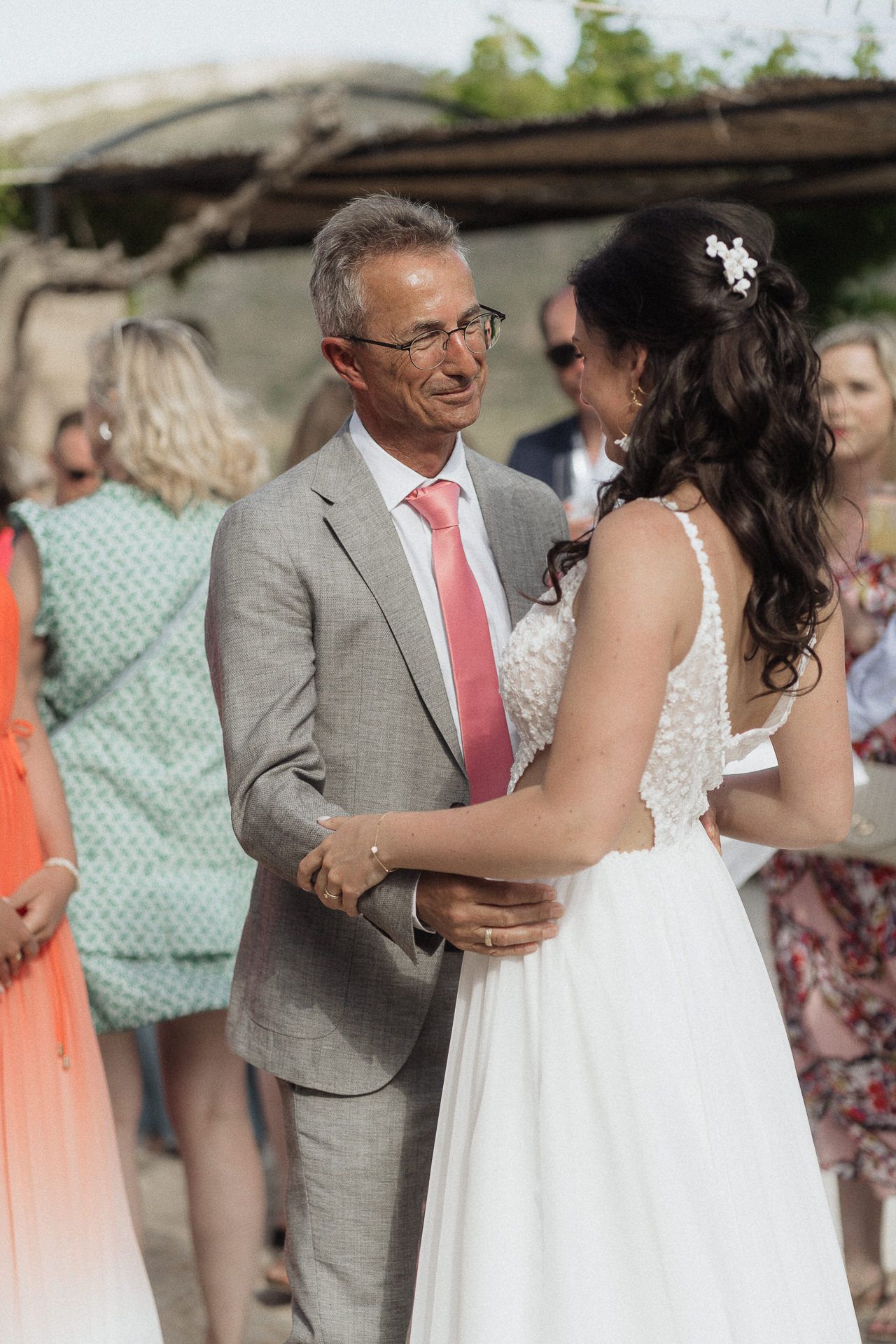 Bride in white gown shares first dance with groom in gray suit and pink tie at outdoor wedding ceremony.