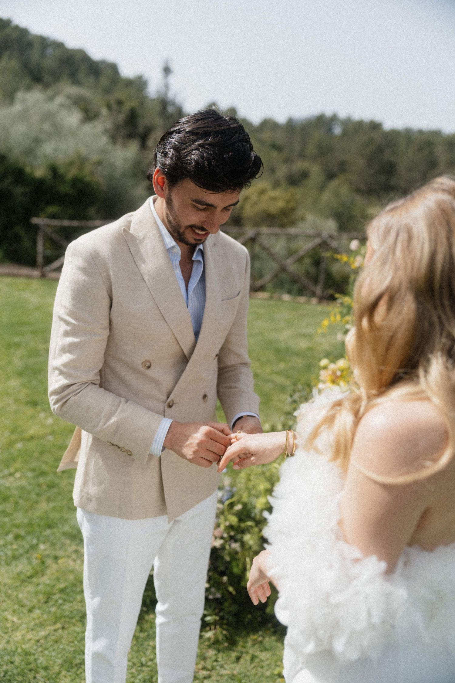 Groom placing wedding ring on brides hand during outdoor ceremony in natural setting.