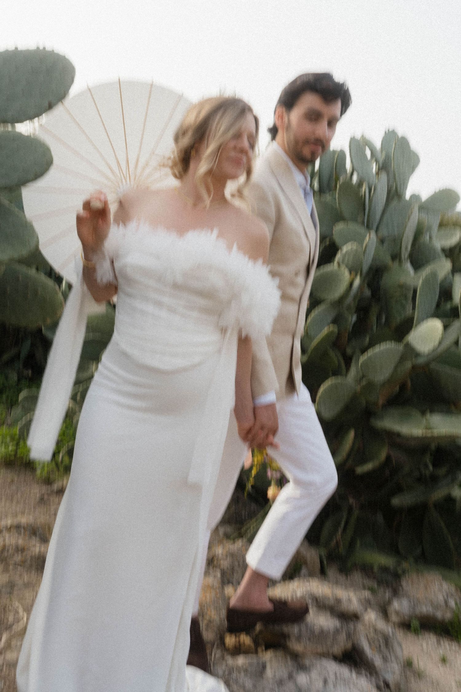 Bride in white feather strapless gown holding parasol walks with groom in beige suit near cactus garden.