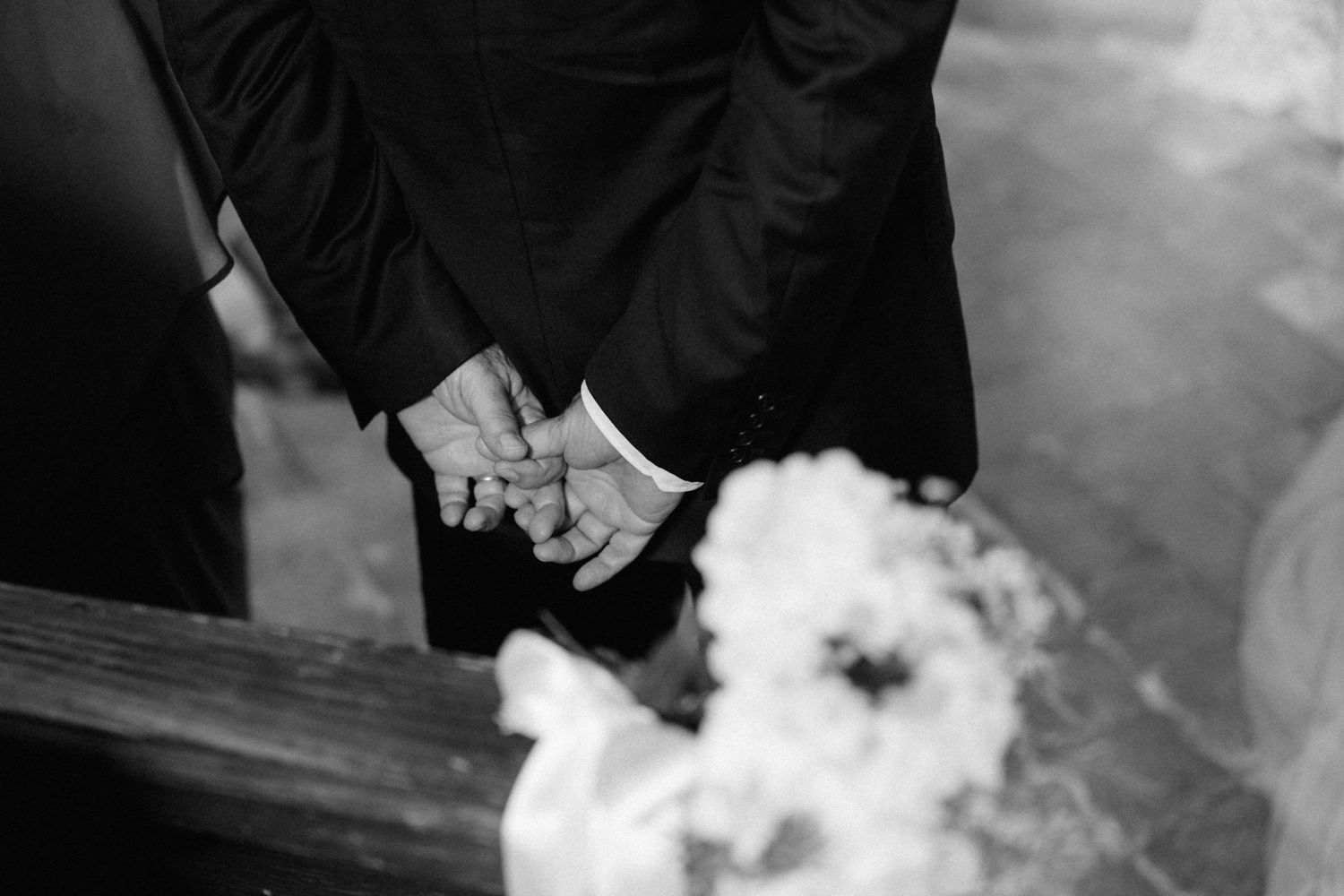 Black and white wedding photo of groom clasping hands, with bridal bouquet in foreground.