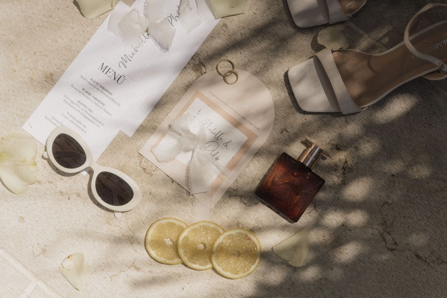 Flat lay of sunglasses, heels, perfume, lemon slices, and papers on sandy surface with shadows.